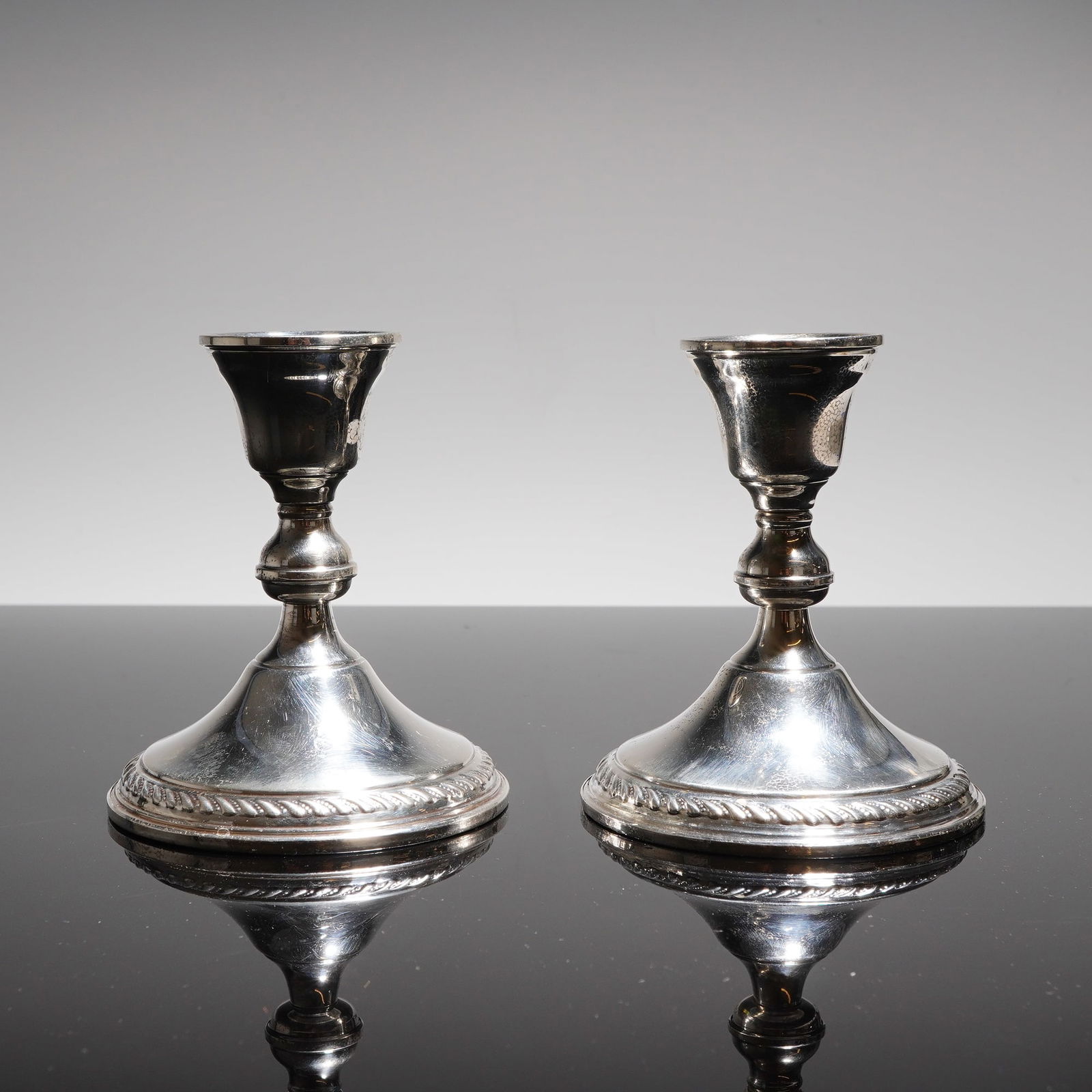 A Pair of Preisner Sterling Weighted Candlesticks Model 724: A pair of marked Preisner sterling weighted candlesticks, model 724. The items feature a traditional design characterized by a wide, spreading circular base decorated with a twisted rope or braid bord