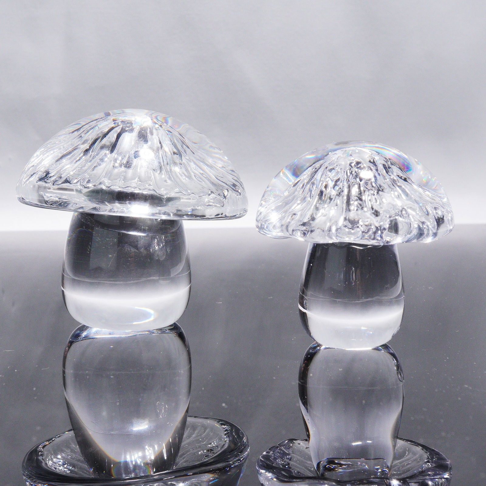 A Pair of Daum France Crystal Mushroom Paperweights: A pair of Daum France clear colorless crystal decorative objects, stylized as mushrooms. Each piece features a dome-shaped cap with a textured, faceted interior, resting on a smooth, tapering cylindri