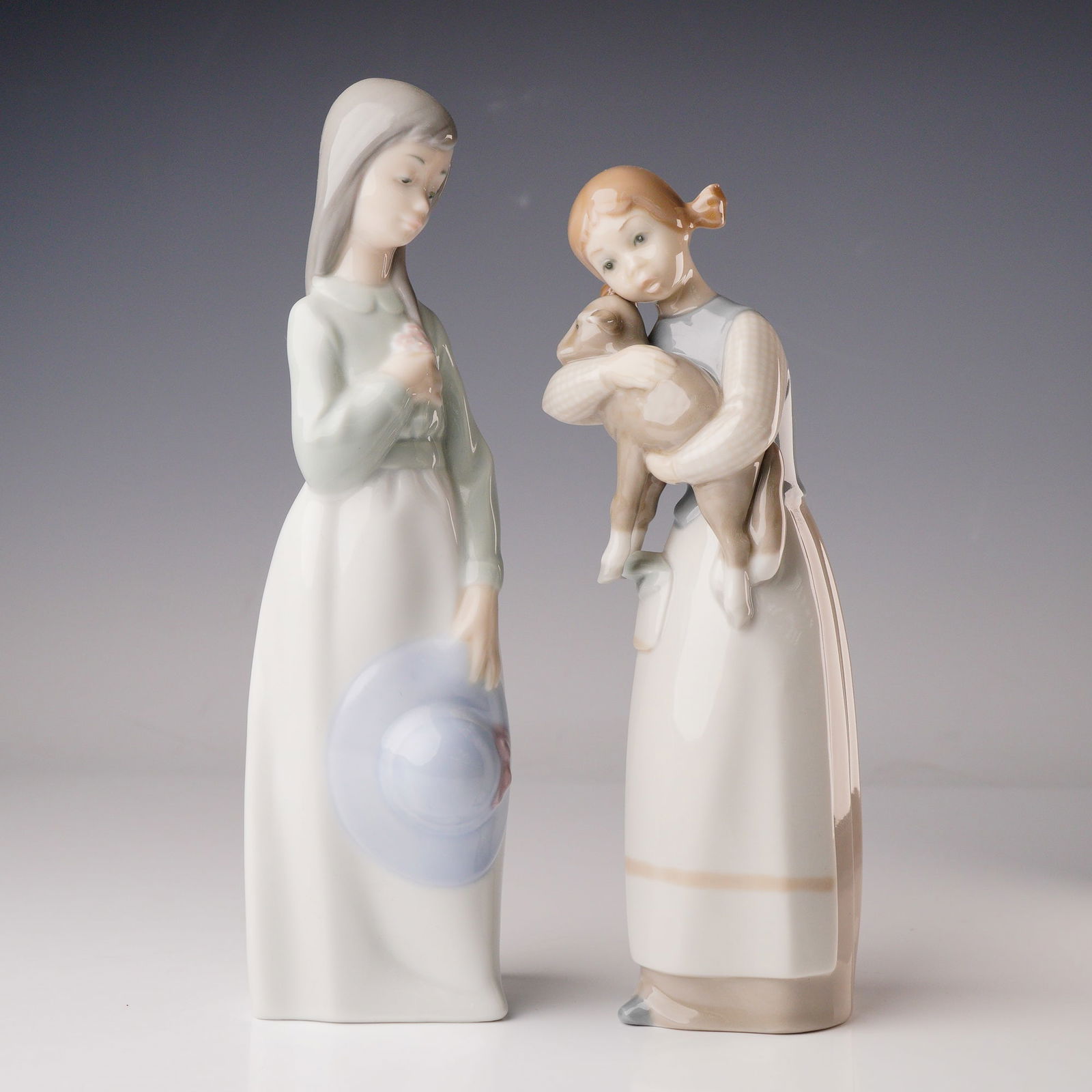 2pc Nao and Lladro Porcelain Figurines: A pair of Spanish glazed porcelain figurines, executed in a contemporary, naturalistic style, depicting standing female figures. Nao figurine features a woman with long greyish-brown hair wearing a wh