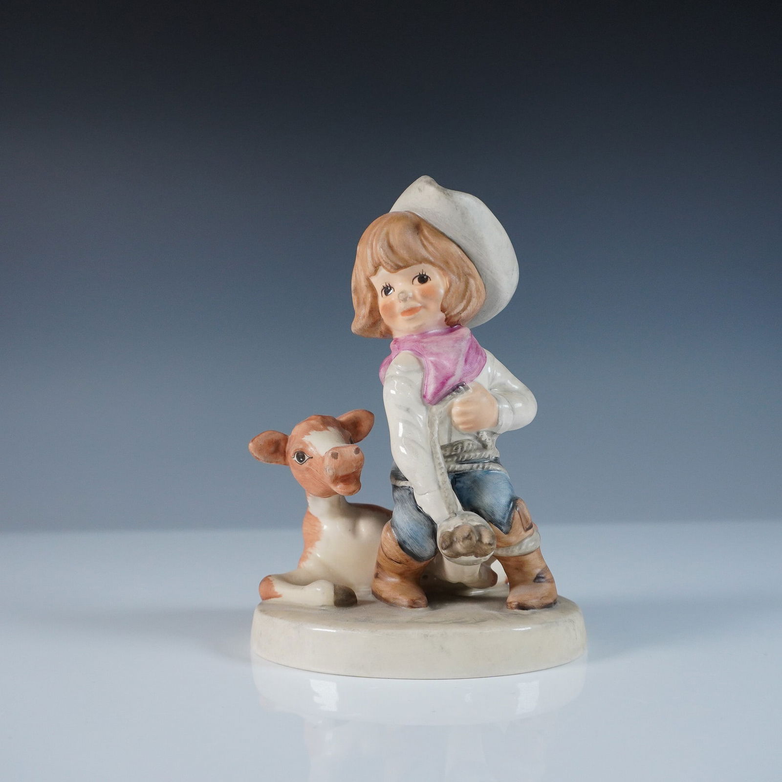 Goebel Figurine, Cowgirl Rodeo: This Goebel figurine, titled Cowgirl Rodeo, captures a charming Western-themed scene of a young cowgirl kneeling beside a cow, dressed in a white cowboy hat, pink bandana, and denim jeans. The figurin