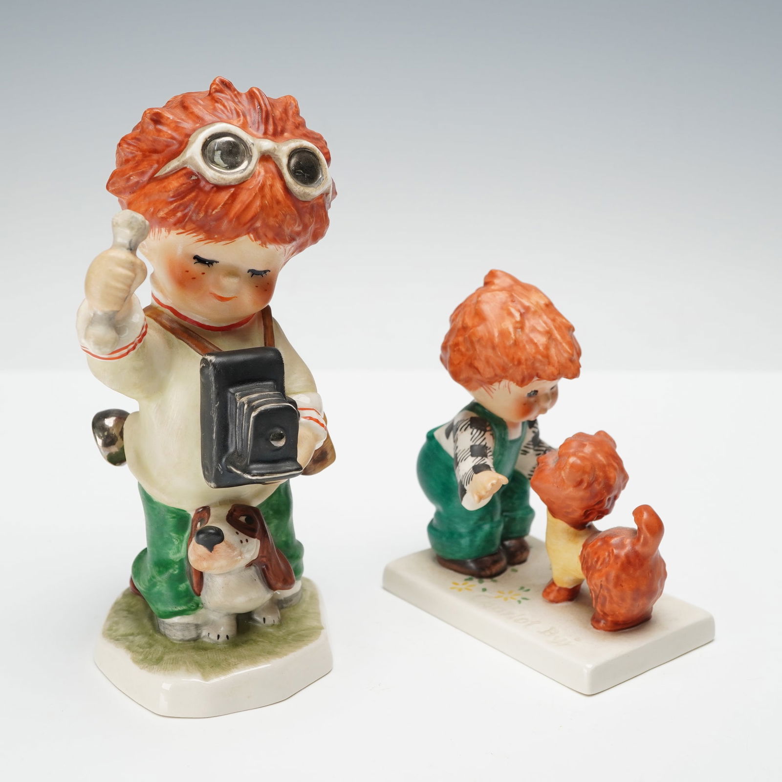 2pc Goebel Red Heads Figurines, Boys with Camera & Dog: Figurines designed by artist Charlot Byj, each depicting an endearing red-haired child with playful expression and bright coloring. One features a young boy holding a camera with his dog at his feet,