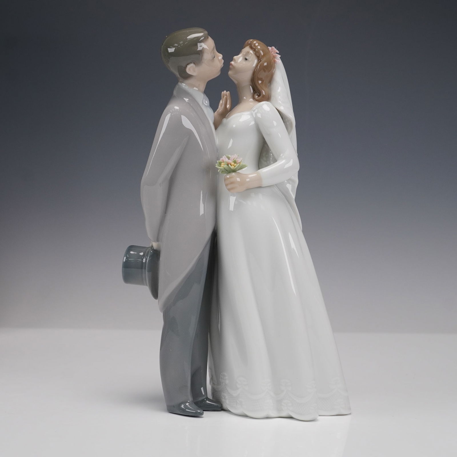 Lladro Porcelain Figurine, A Kiss to Remember 1006620: Lladro porcelain figurine titled A Kiss to Remember, number 1006620, depicting a romantic wedding scene with a bride and groom in soft pastel glazes. The groom wears a grey morning coat and holds his