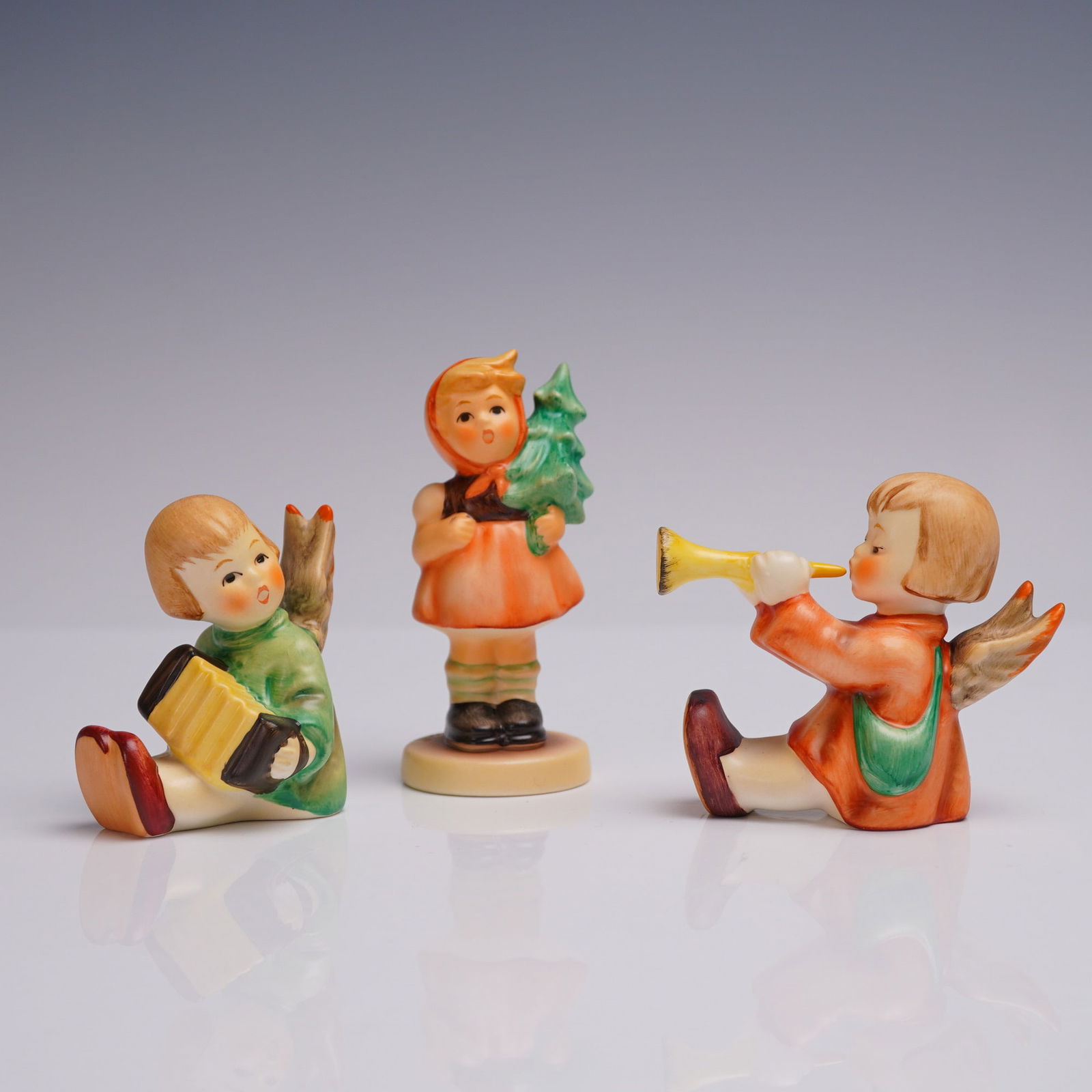 3pc Goebel Porcelain Figurines - Girl with Fir Tree and Two Angels: A charming trio of hand-painted Goebel porcelain figurines from Germany, each marked with blue Goebel backstamps and dated 1998. This lot includes a girl in an orange dress holding a fir tree (model 2