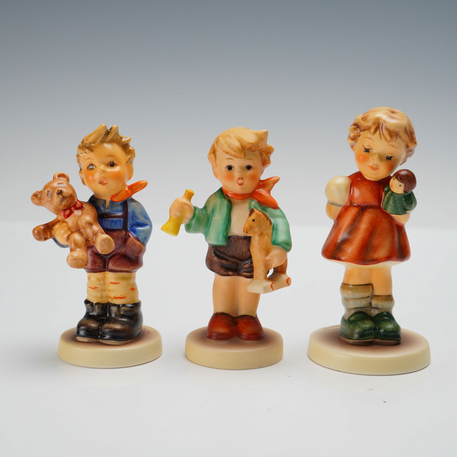 3pc Goebel Hummel Figurines, Childhood Playtime Series: Charming grouping of three Goebel Hummel porcelain figurines, each depicting a child with a beloved toy. Includes Teddy Bear Boy 2074/A, holding a teddy bear; Boy with Horse 239, with a pull toy horse