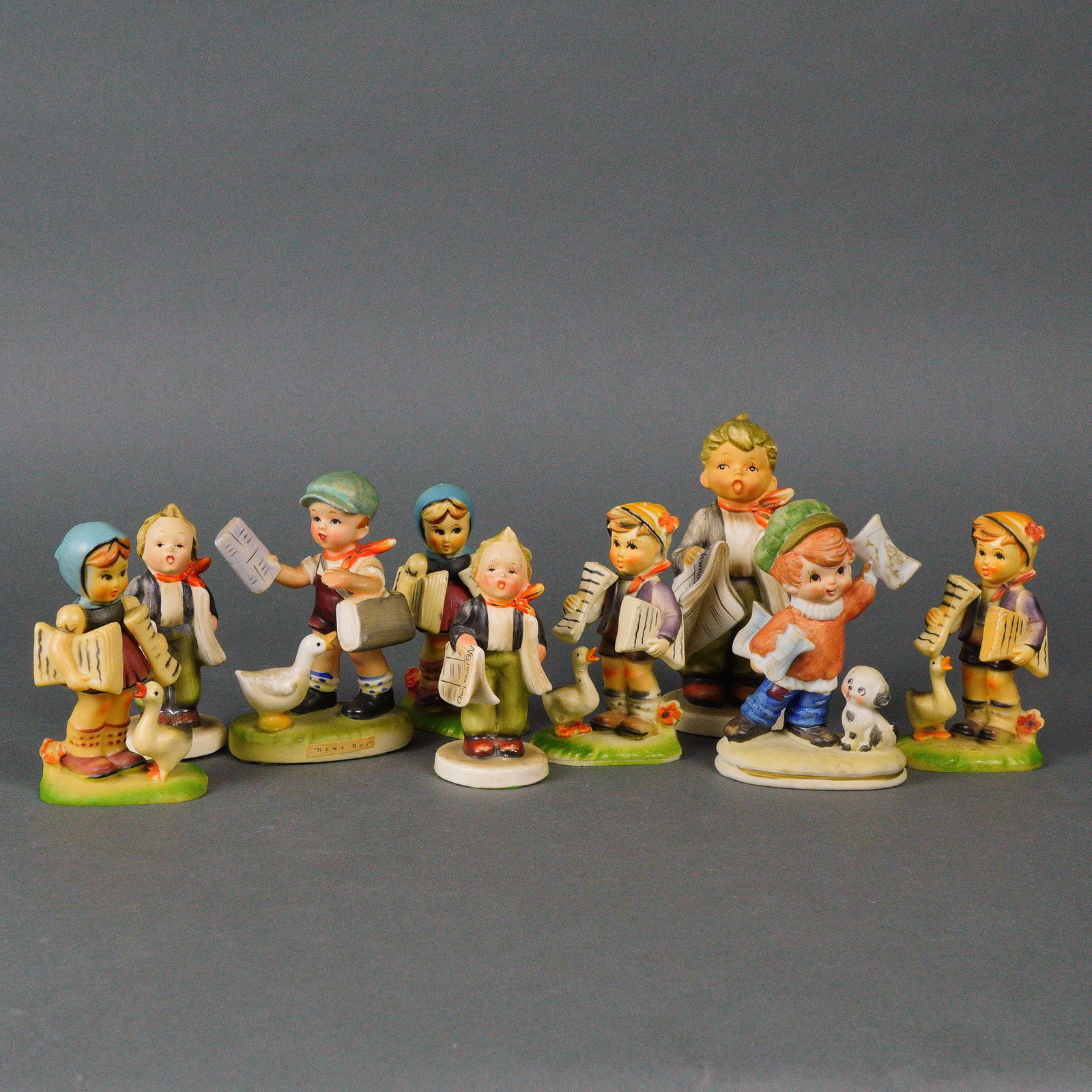A Collection of Eleven Hummel Goebel Figurines: A collection of eleven hand-painted glazed ceramic figurines by Hummel Goebel, primarily depicting rosy-cheeked, cherubic children engaged in various activities such as playing instruments, carrying n