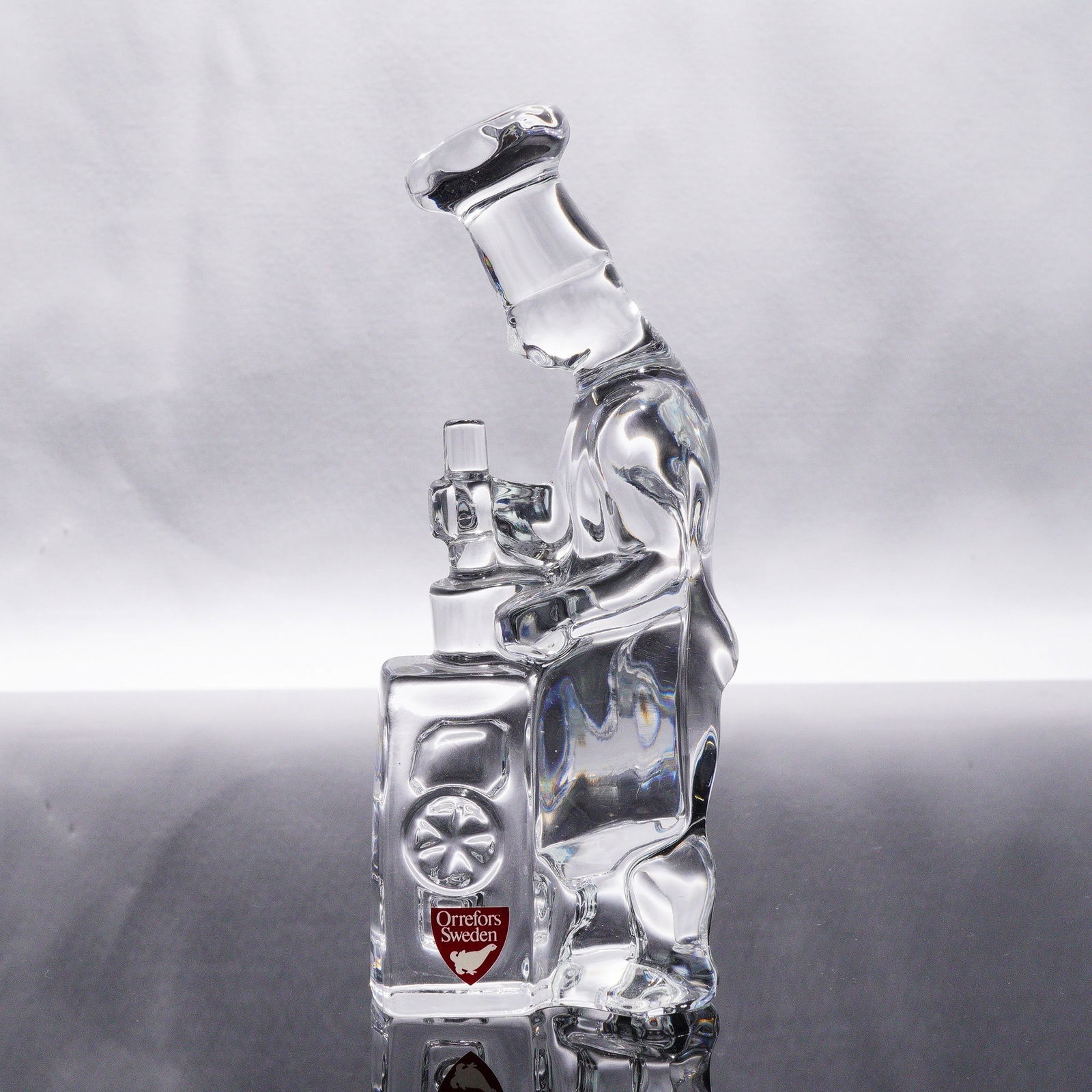 Orrefors Glass Figure - 'The Chef' by Olle Alberius, Craftsmen Series: A clear, colorless glass sculptural decorative item by Orrefors Sweden, designed by Olle Alberius, depicting a stylized chef figure operating a piece of kitchen equipment, possibly a grinder or mixer.