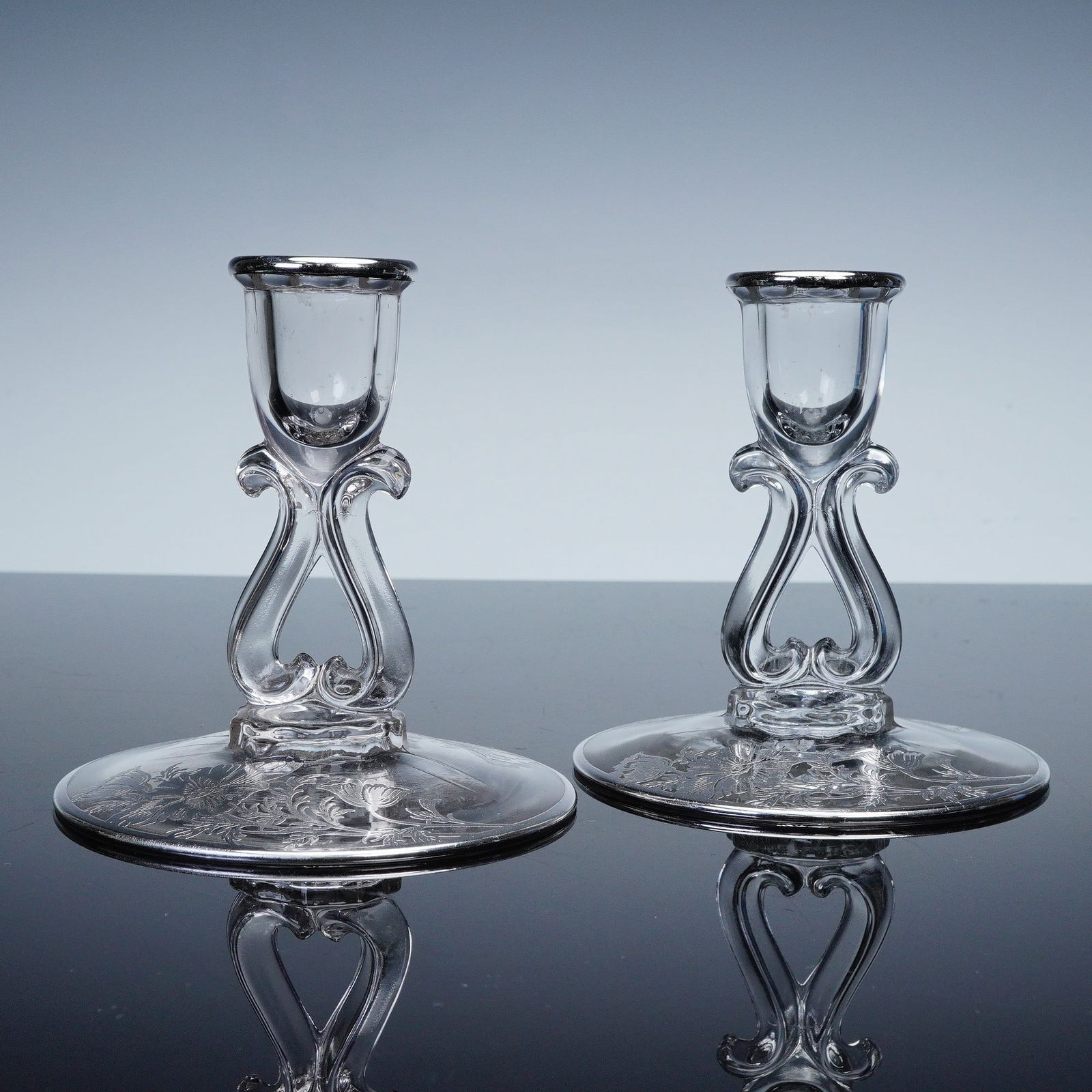 2pc Silver Toned Floral Glass Candlesticks: A pair of elegant glass candlesticks featuring scroll shaped stems and silver toned rims. The wide bases are decorated with intricate etched floral designs, creating a refined and decorative presentat