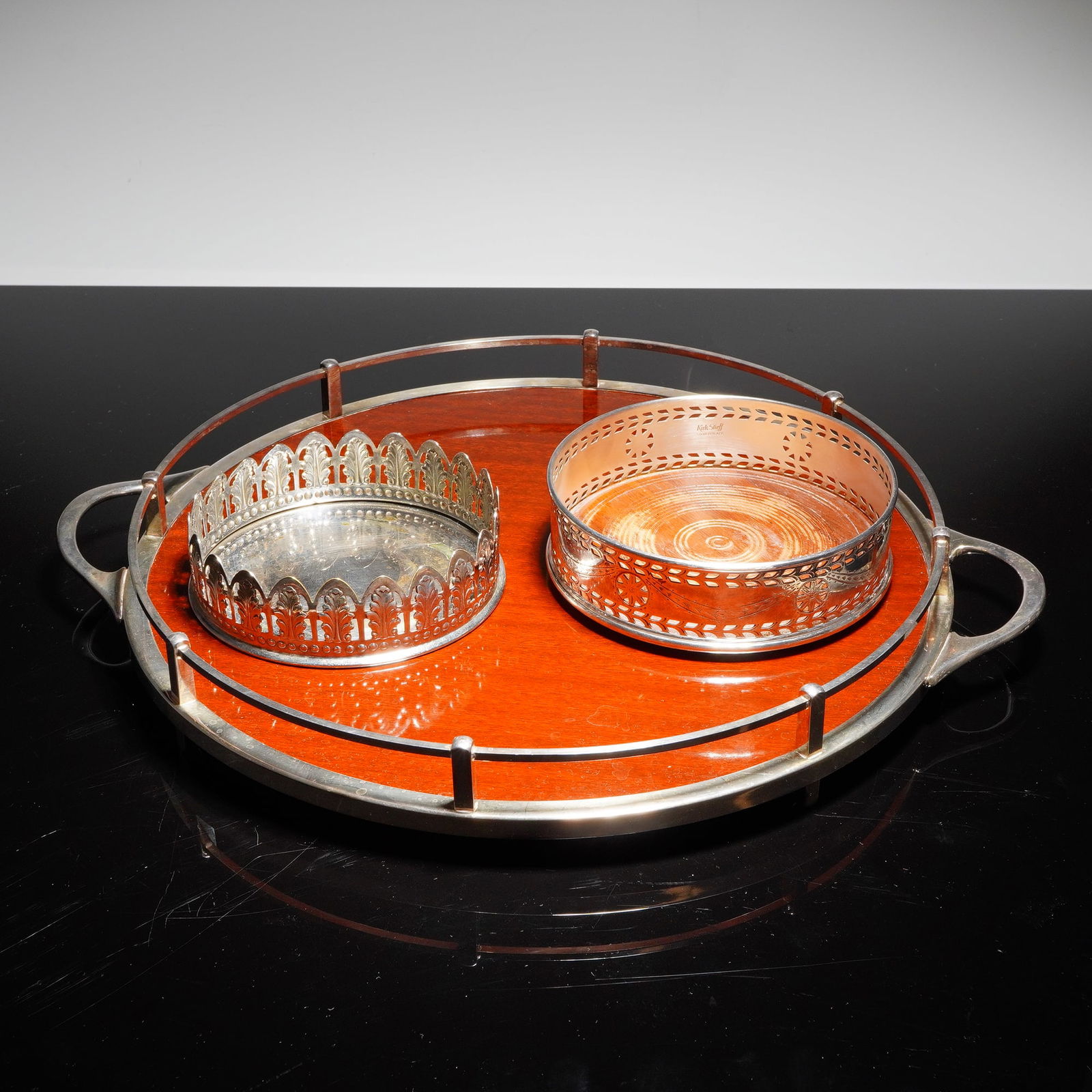 A Trio of Serving Wares: Wolff Formica Tray & Two Silverplate Wine: A collection of three serving items consisting of one oval gallery tray and two separate silverplate wine bottle coasters. The tray features a highly polished reddish laminate surface, identified by a