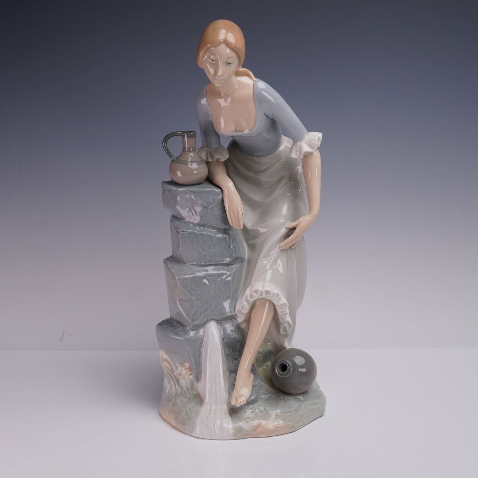 Nao by Lladro Porcelain Figurine, Girl from the Fountain: Nao by Lladro porcelain figurine titled Girl from the Fountain, designed in Spain. This charming figurine depicts a young woman resting by a stone fountain with a small jug at her side and a larger ve