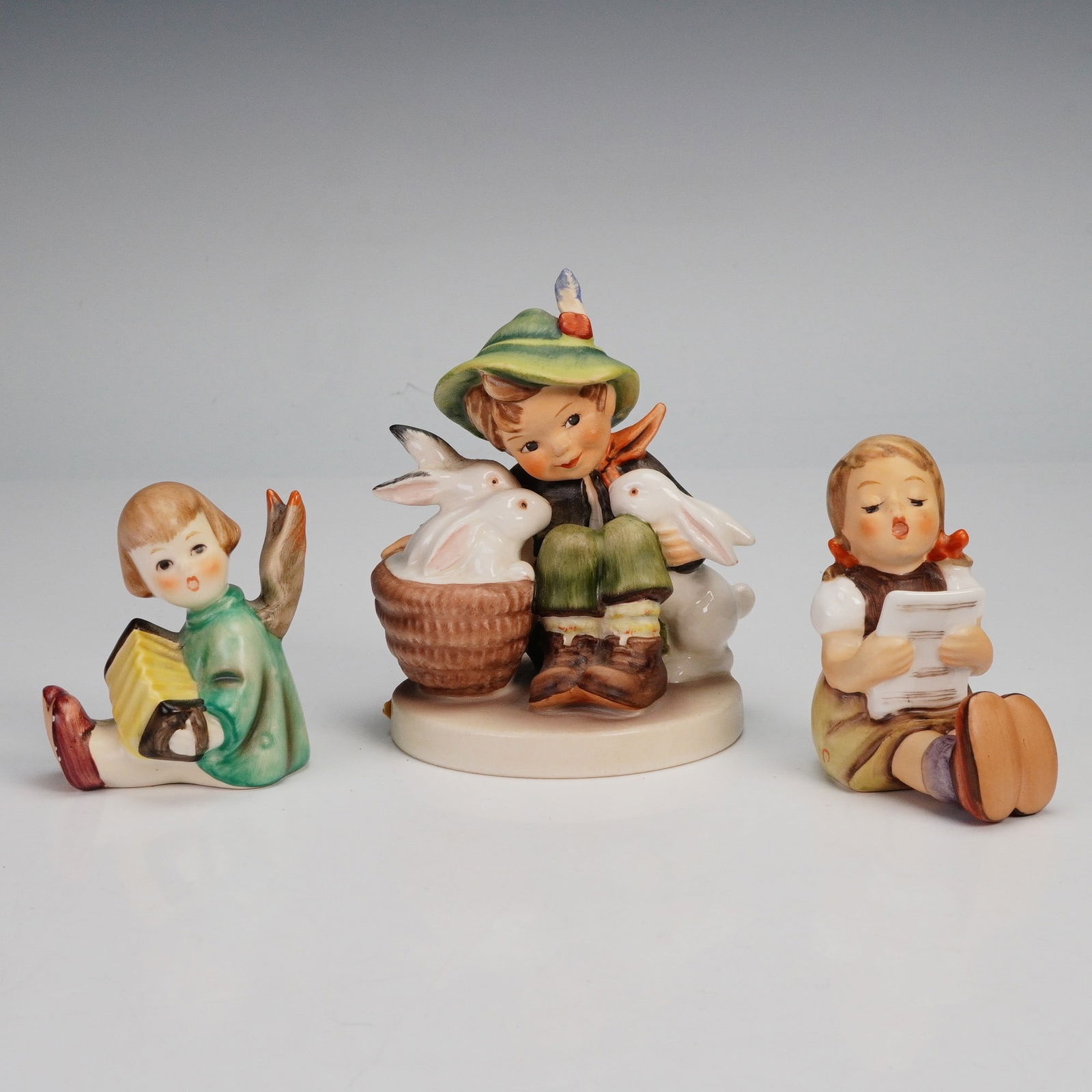 3pc Goebel Hummel Figurines, Playmates, Music and Angel: A group of three Hummel porcelain figurines including Playmates featuring a boy with two white rabbits, Girl with Sheet of Music depicting a young girl singing, and Angel with Accordion. Each piece is