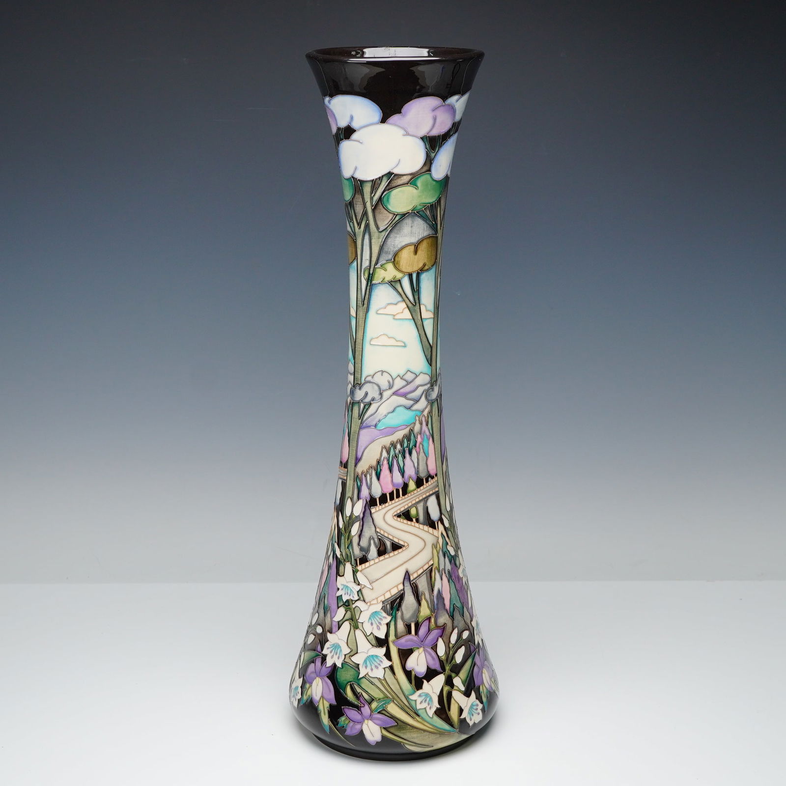 Moorcroft by Nicola Slaney Pottery Vase, Blue Ridge Parkway: Designed in a tall tapered form, this Moorcroft vase features the Blue Ridge Parkway scene rendered with stylized trees, soft pastel clouds, and a winding mountain road bordered by floral motifs. The