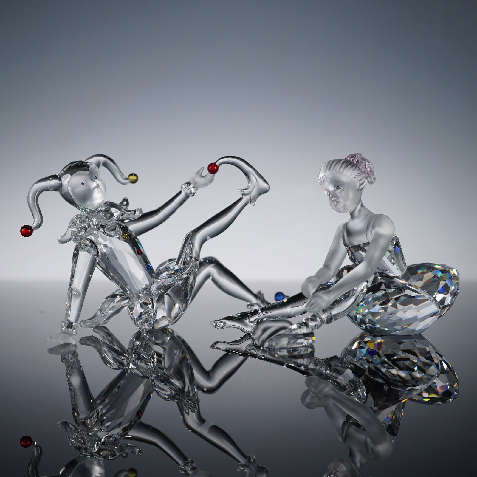 A Pair of Swarovski Crystal Jester and Ballerina Figurines: A pair of Swarovski crystal figurines, identified by the etched Swarovski Swan Logo and registered mark visible. The set includes two pieces: a Jester and a Ballerina. The Jester figurine is construct