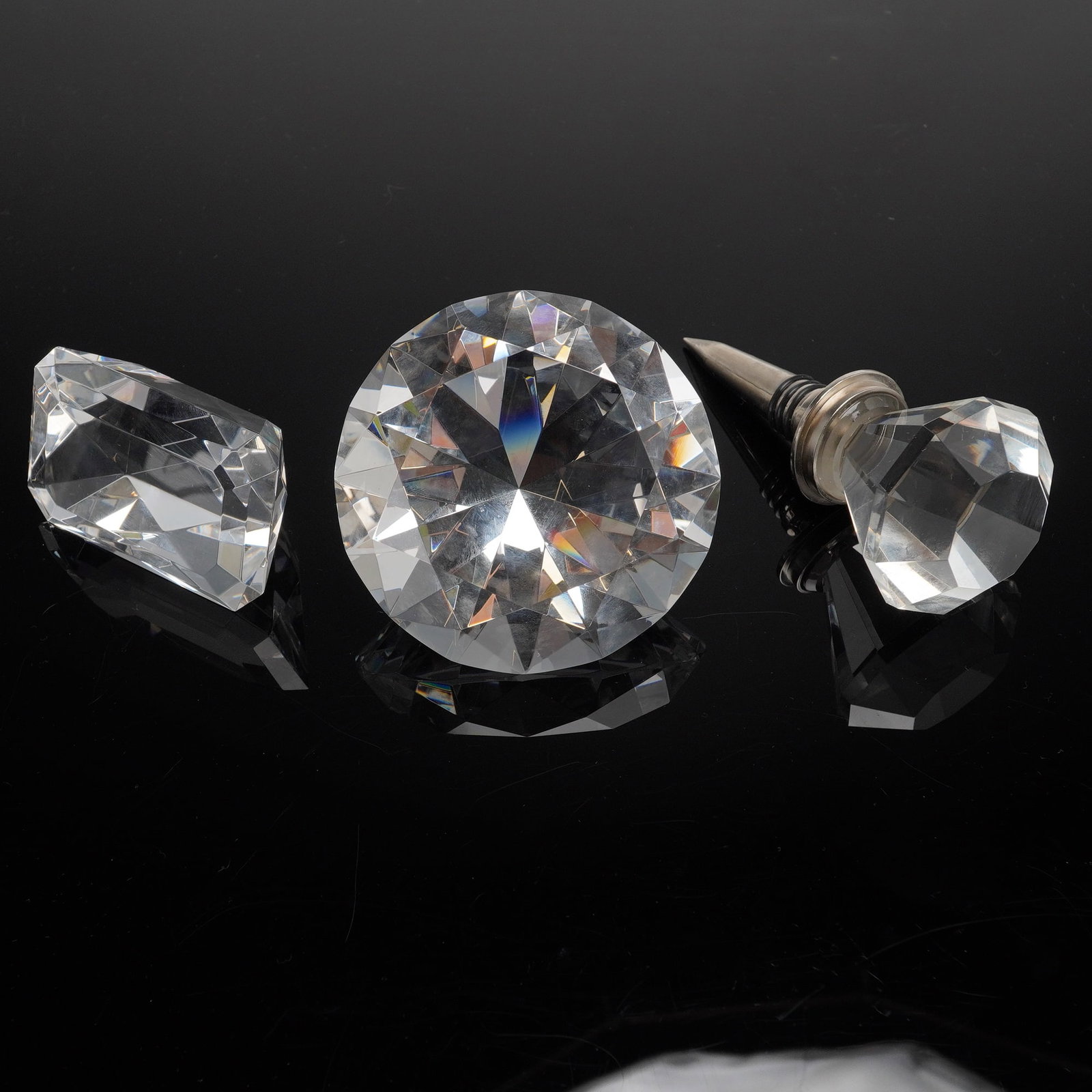 A Trio of Clear Faceted Crystal DiamondPaperweights and Bottle Stopper: A trio of decorative objects crafted from clear, highly faceted crystal or heavy glass, designed to simulate large cut gemstones. The lot includes two large display objects intended as paperweights an