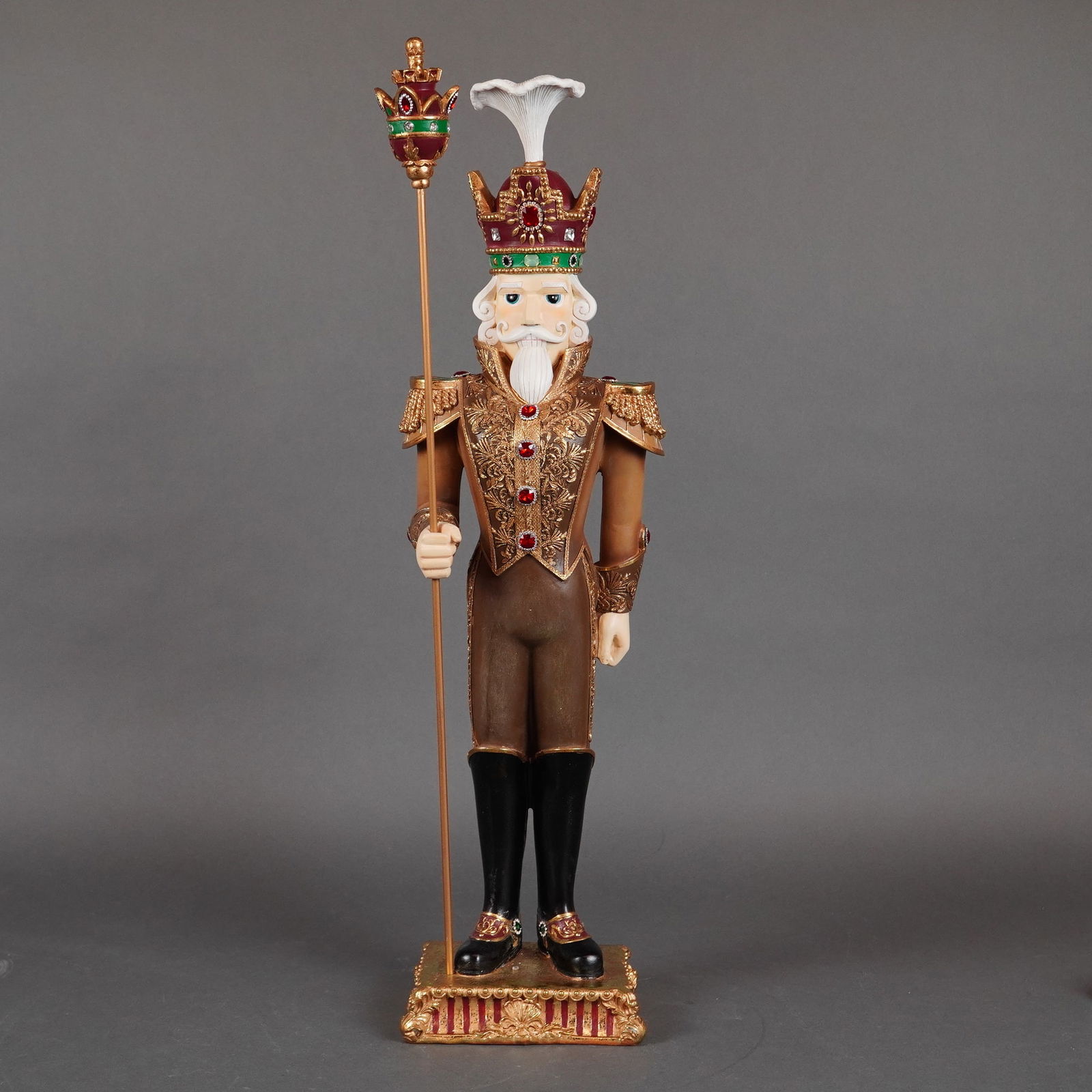 K&K Interiors Gold and Burgundy Jeweled Resin Soldier: A tall, decorative soldier , rendered in a richly embellished palette of brown, gold, red, and green tones, and standing upright upon a molded rectangular base with gilt accents and striped detailing.