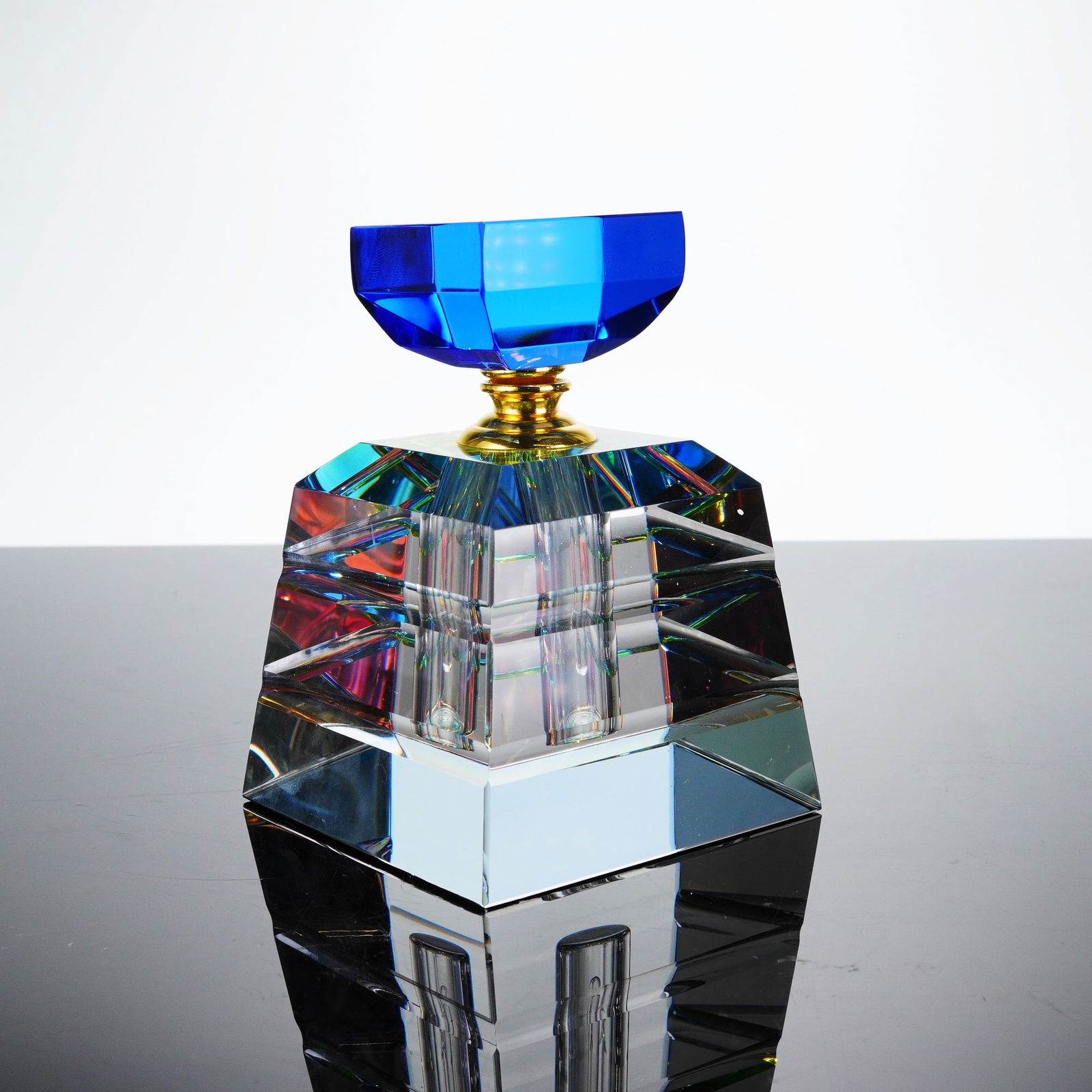 A Crystal Perfume Bottle with Stopper: A single modern perfume bottle, constructed from heavy clear faceted crystal. The body features a substantial, geometric, trapezoidal base designed to maximize light refraction. The neck is fitted wit