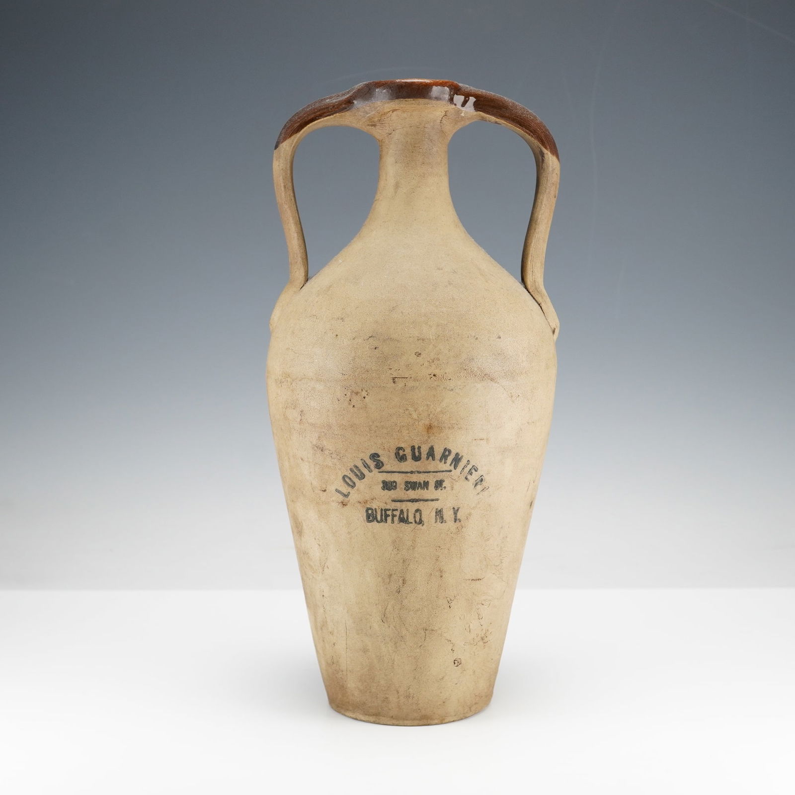 Antique Louis Guarnieri 389 Swan St. Buffalo, NY Stoneware Wine Jug: Two handled stoneware advertising wine jug. This tall vessel features a narrow neck and a small opening. The majority of its body displays a light brown finish with a contrasting dark brown glaze on t