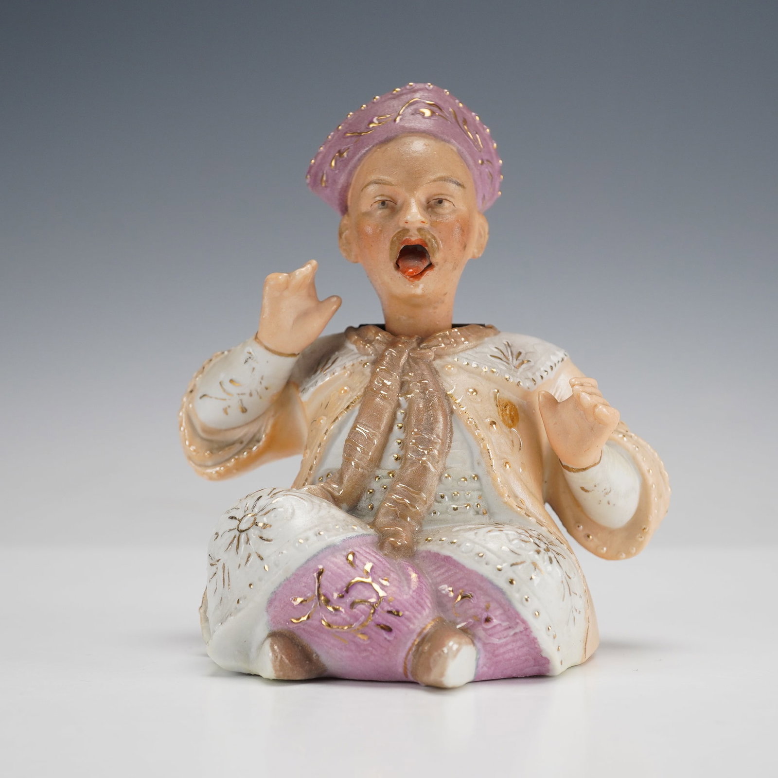 Continental Bisque Porcelain Nodding Figure: The seated figure wears a lavender cap and a light-colored robe over patterned trousers, all extensively adorned with gold gilding in floral and starburst motifs. He is rendered with a caricatured exp