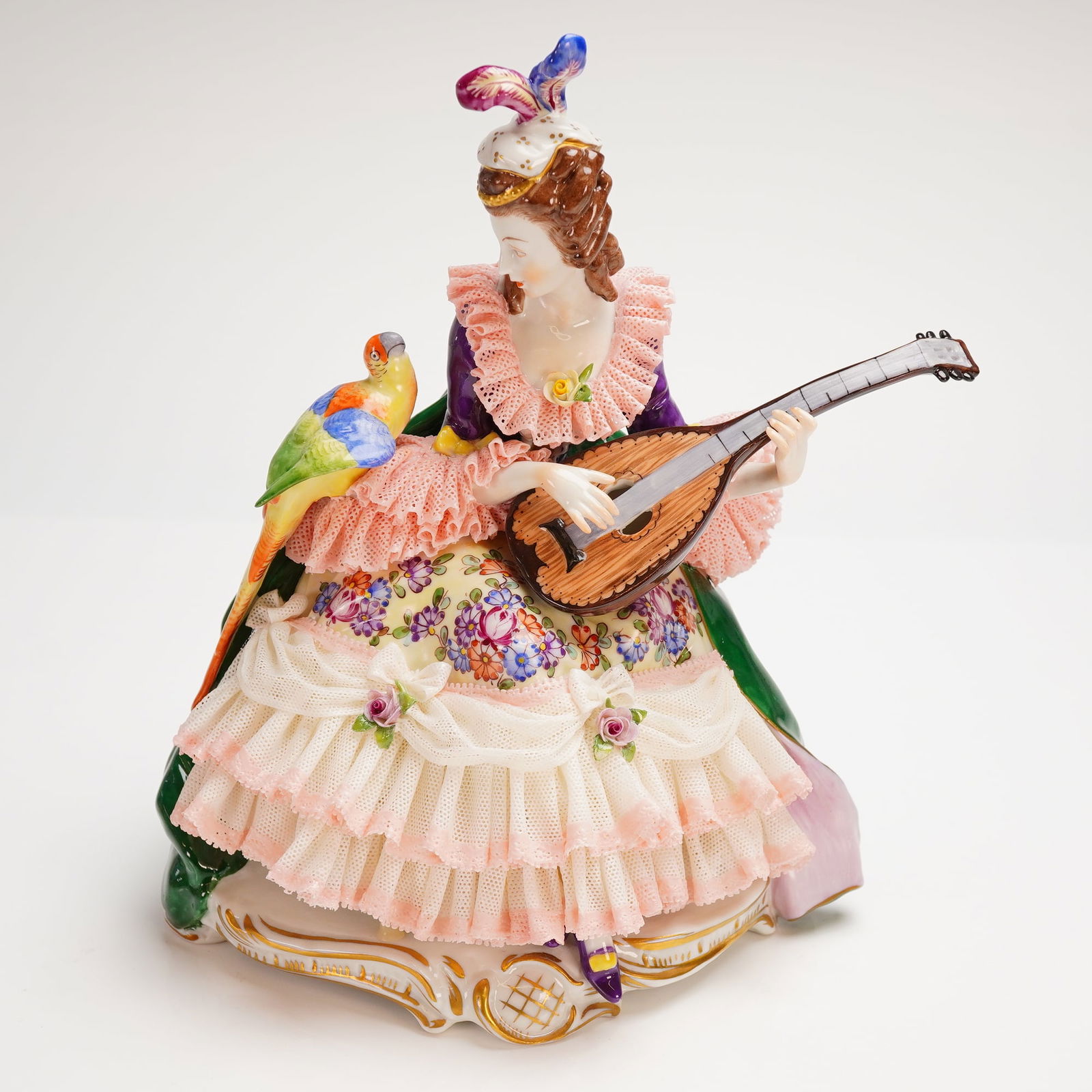 Dresden Porcelain Lace Figurine of a Woman with Parrot Playing a Mandolin: A single polychrome porcelain Lace Figurine crafted in the Rococo-revival style, attributed to the Dresden school of manufacture. The figurine depicts a noblewoman seated on an ornate, gilt-accented b