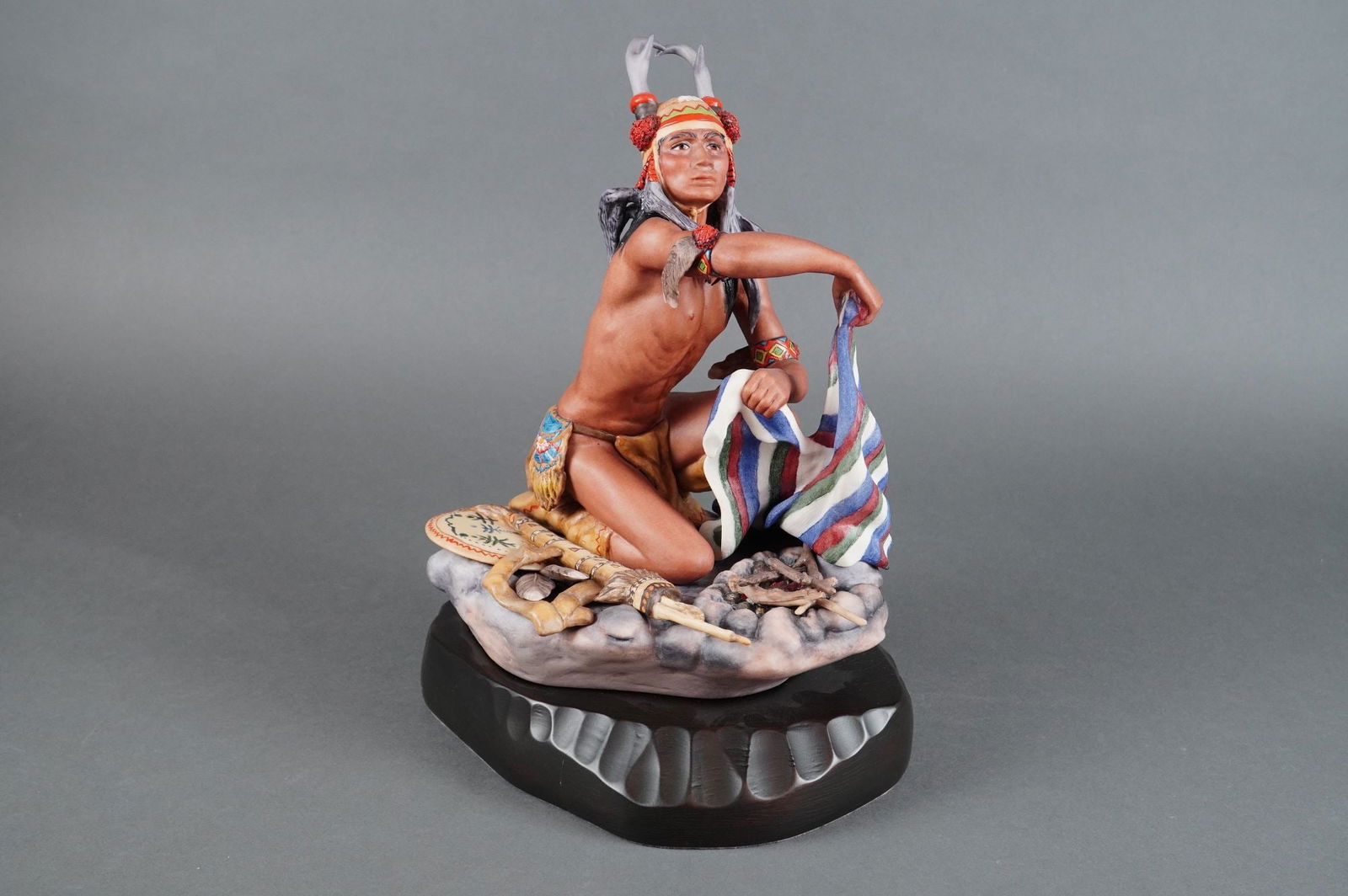 Cybis Porcelain Figurine Apache Chato: A single Cybis porcelain figurine, titled "Apache Chato," depicting a kneeling indigenous male figure in elaborate ceremonial attire. The figurine is executed in matte bisque porcelain with detailed p