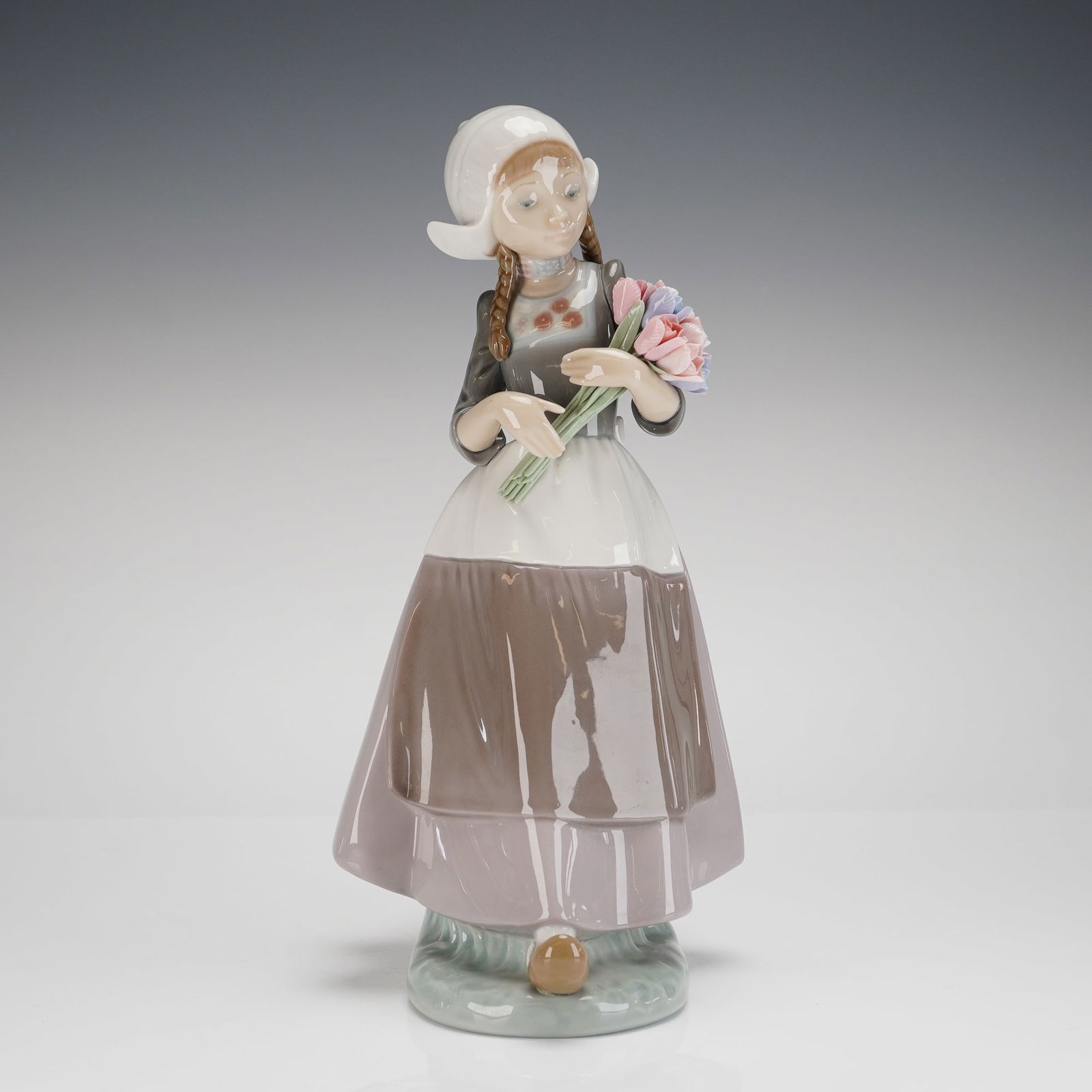 Lladro Dutch Girl Porcelain Figurine: A single glazed porcelain figurine by Lladro, depicting a young girl in traditional Dutch or stylized folk costume. The figure wears a white bonnet, a gray jacket with floral details, and a mauve apro