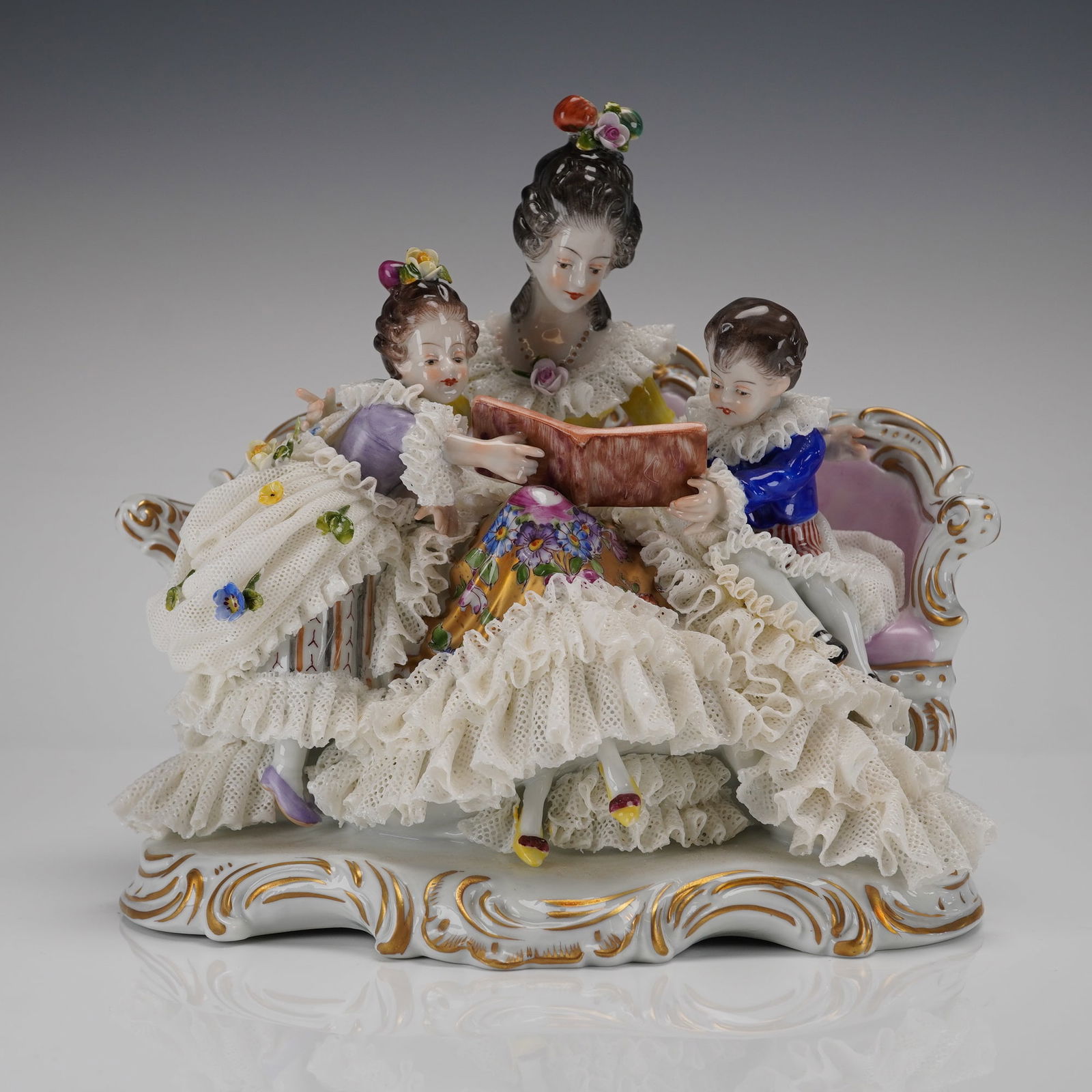 Dresden Porcelain Lace Figurine Mother and Children Reading: A Dresden Porcelain Lace Figurine depicting a mother and children reading, executed in polychrome-painted and gilt porcelain in the Rococo Revival style. This single figural group sculpture, often mar