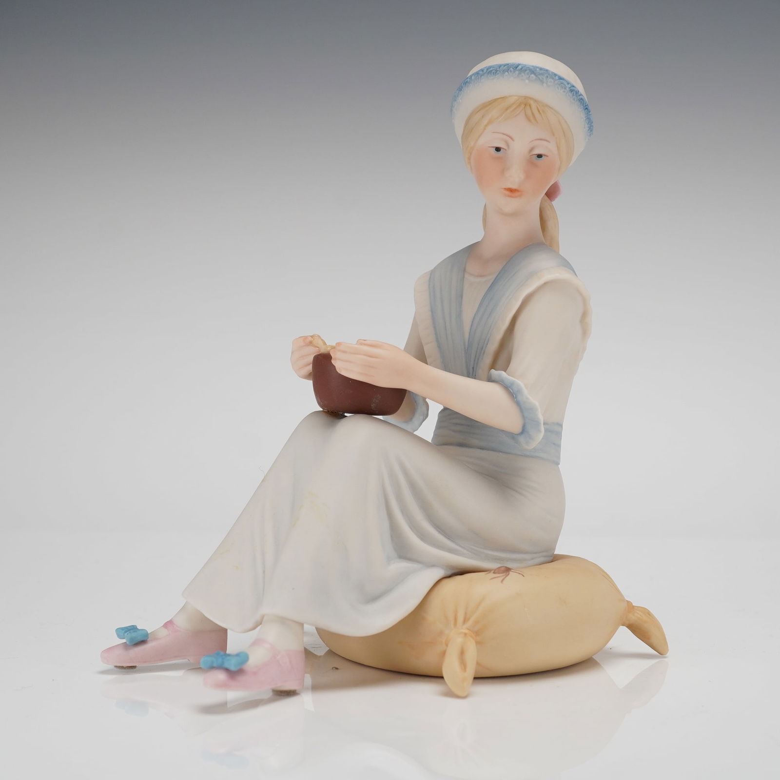 Cybis Porcelain Figurine Little Miss Muffet: A Cybis Figurine, executed in bisque (matte) porcelain, depicting the subject Little Miss Muffet. The figure is modeled as a young woman seated upon a pale yellow cushion, wearing a long cream-colored