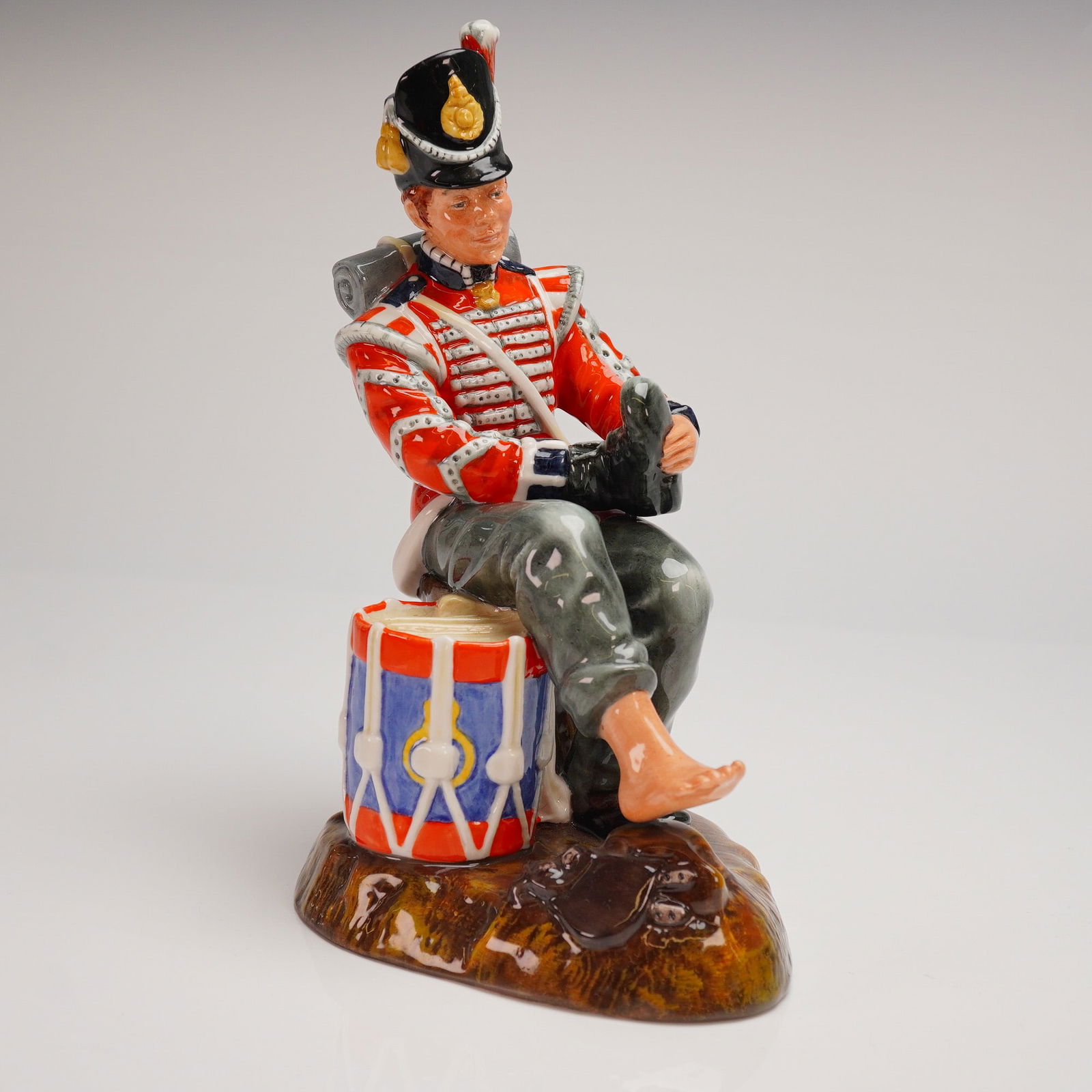 Royal Doulton Glazed Porcelain Figurine Drummer Boy HN 2655: A single Royal Doulton glazed porcelain figurine titled Drummer Boy. The figurine depicts a historic uniformed soldier, likely a Grenadier or similar regiment, seated upon a military drum rendered in