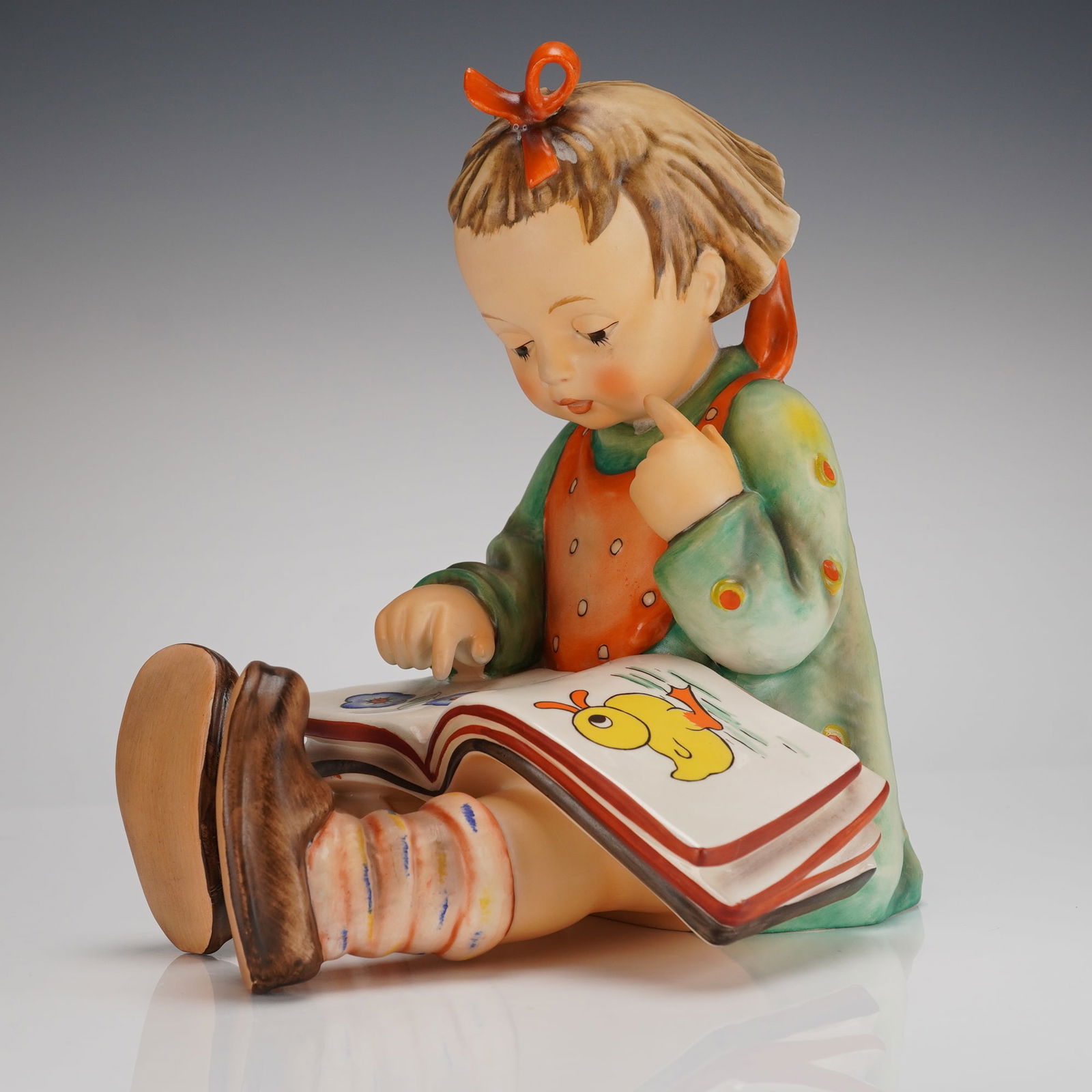 Goebel M.J. Hummel Figurine Bookworm, Hum 3/II: A single glazed ceramic figurine by Goebel, designed by M.J. Hummel, titled Bookworm (Hum 3/II). The hand-painted figurine depicts a young girl seated with her legs crossed, engrossed in reading an op