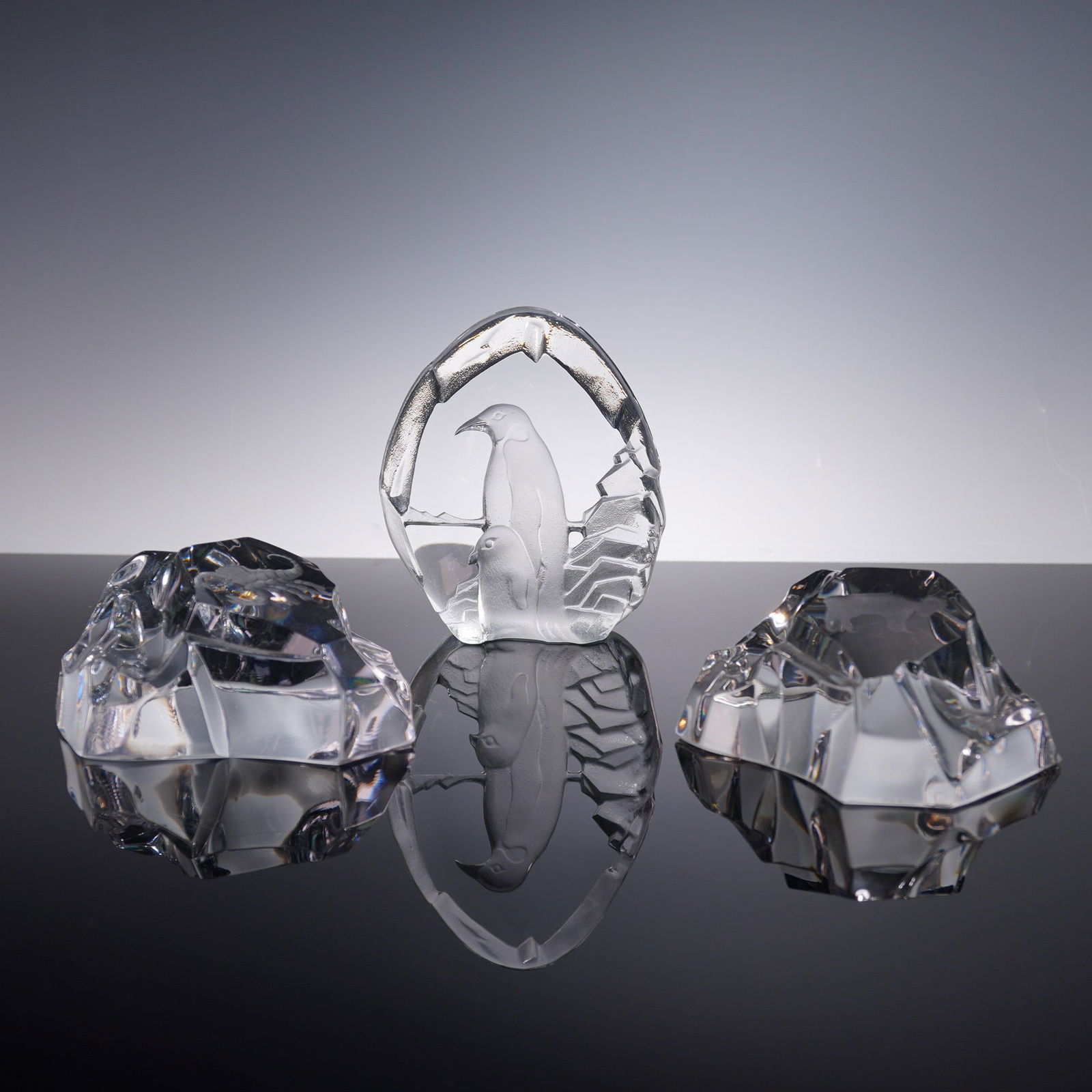 3pc Nybro Crystal Paperweights: Group of three Swedish crystal paperweights by Nybro, comprising a central sculpted panel featuring two penguins nestled within a stylized arched ice formation, flanked by two freeform iceberg-inspire