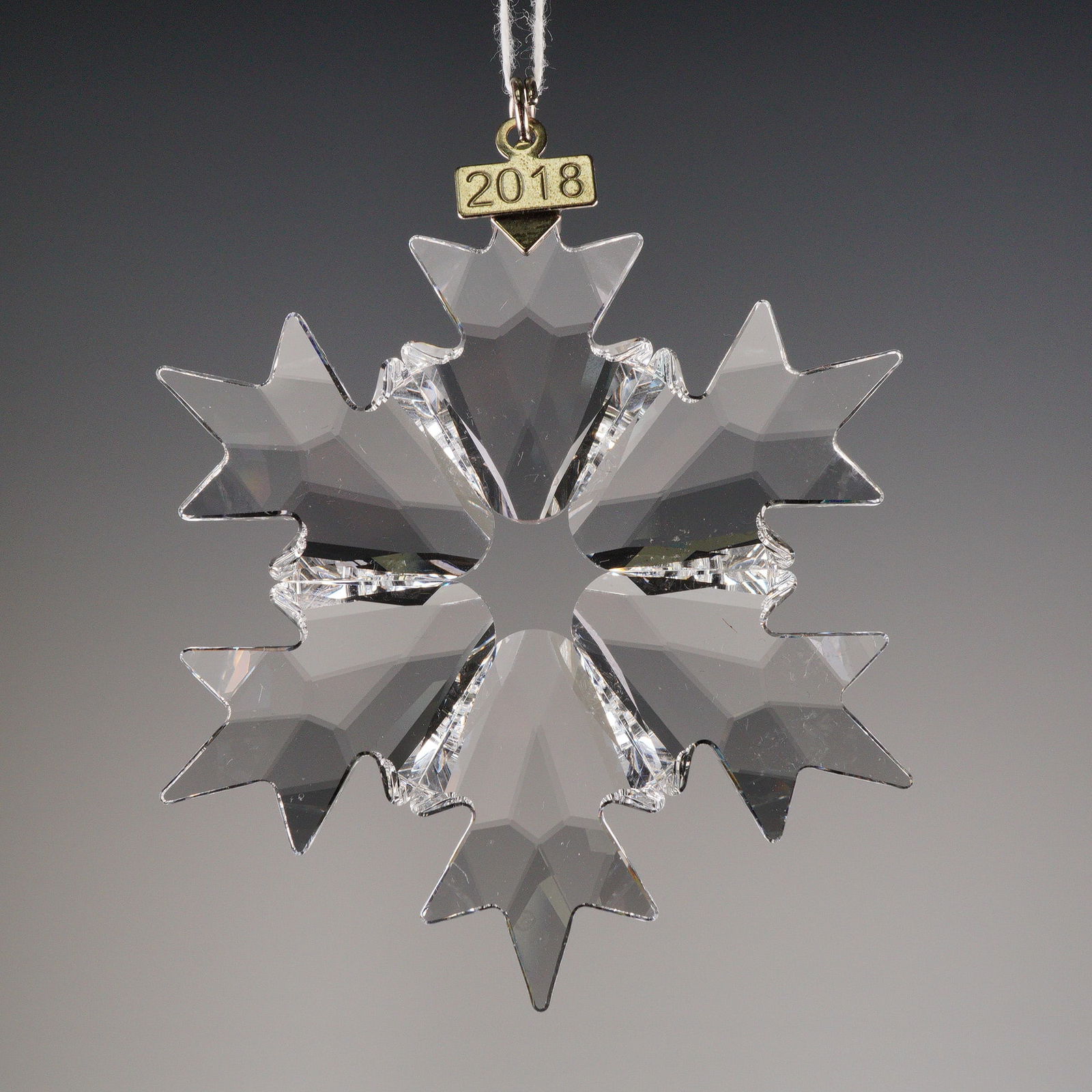 Swarovski 2018 Annual Edition Crystal Snowflake Ornament: A Swarovski 2018 Annual Edition Snowflake. The ornament is crafted from clear, cut crystal in a six-pointed snowflake design. The tag is engraved with the date 2018 on the front. The metal base bears