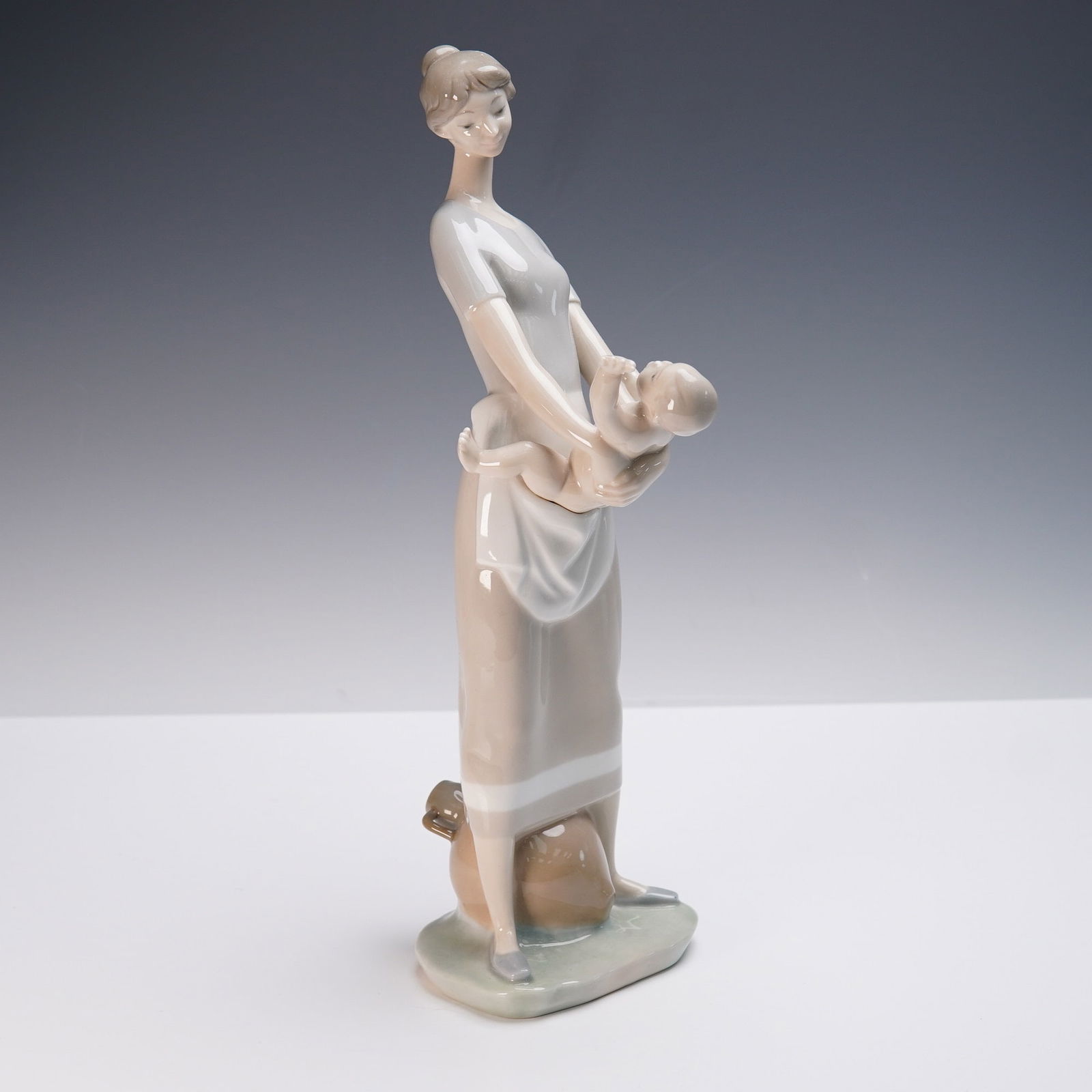 Lladro Porcelain Figurine, Mother and Child 01004575: A touching depiction of maternal love, this Lladro porcelain figurine titled Mother and Child features a serene young woman gently cradling an infant in her arms while standing in a relaxed pose. Desi