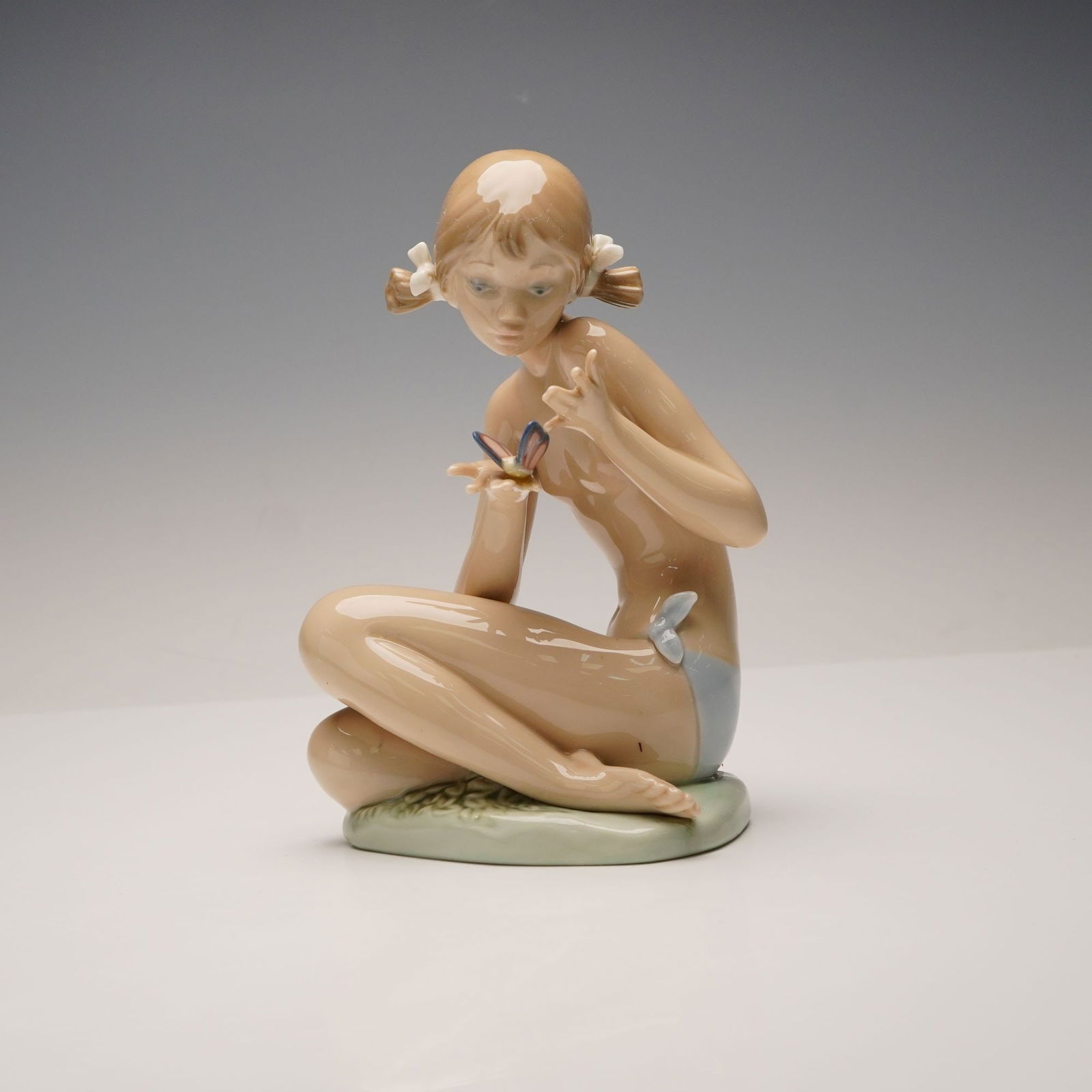 Lladro Porcelain Figurine, Free As A Butterfly 1001483: This glazed porcelain figurine depicts a young girl in a seated, cross-legged pose. She wears light blue briefs and has her hair styled in pigtails adorned with white floral accents, delicately holdin