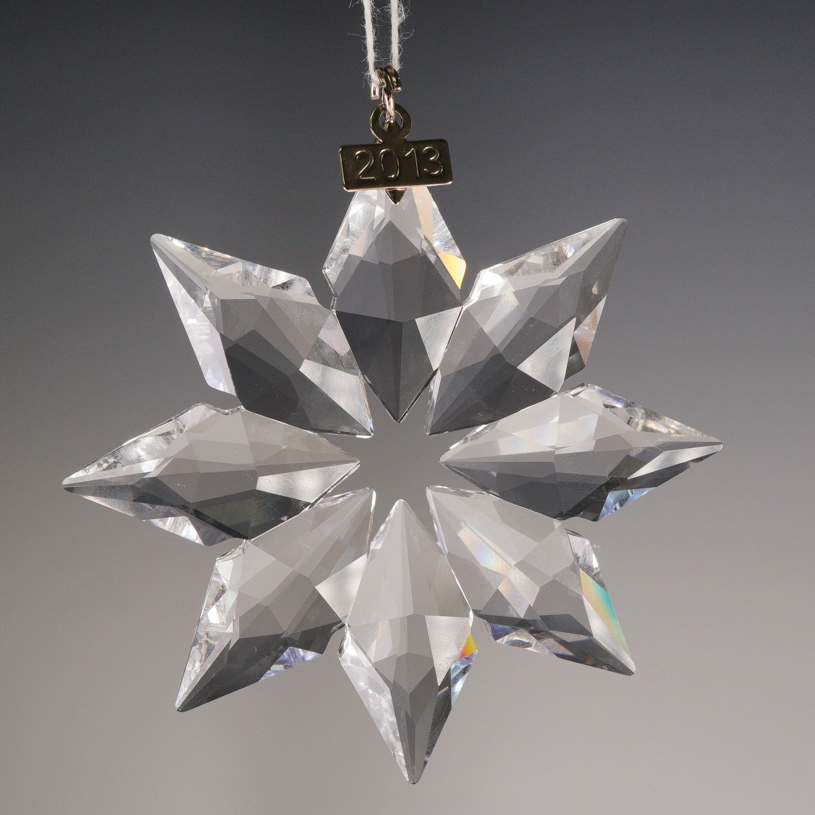Swarovski Annual Edition 2013 Crystal Snowflake Ornament: The piece is fashioned as an eight-pointed snowflake, composed of clear faceted crystal designed for light refraction. The silver toned tag is engraved with the year "2013." The suspension clip itself