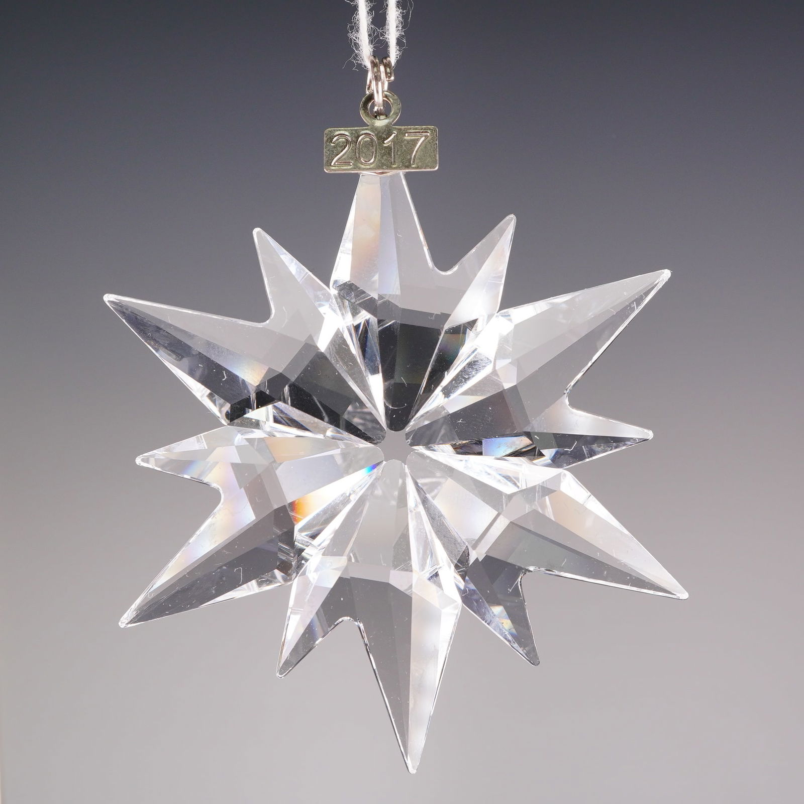 Swarovski Crystal 2017 Annual Edition Snowflake Ornament: The piece is crafted from clear faceted crystal in a geometric, twelve-pointed snowflake design. The silver toned tag is engraved with the year 2017. The Swarovski logo is present on the metal compone