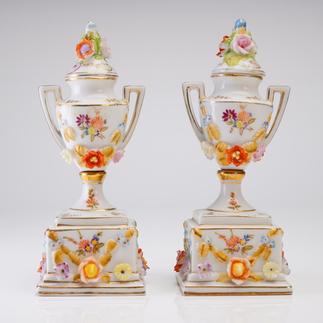 A Pair of Dresden Style Porcelain Floral Encrusted Lidded Miniature Urns: A pair of Dresden style porcelain lidded miniature urns, likely Continental European production, dating to the late 19th or early 20th century. Each item is crafted from white glazed porcelain in a co