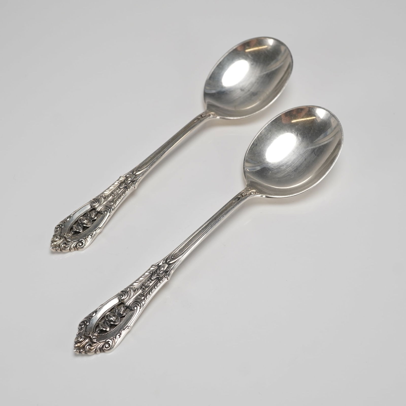 A Pair of Wallace Rose Point Sterling Silver Cream Soup Spoons: A pair of sterling silver soup spoons manufactured by Wallace Silversmiths in the highly recognized Rose Point pattern. These cream soup spoons feature highly ornate handles characteristic of the Baro
