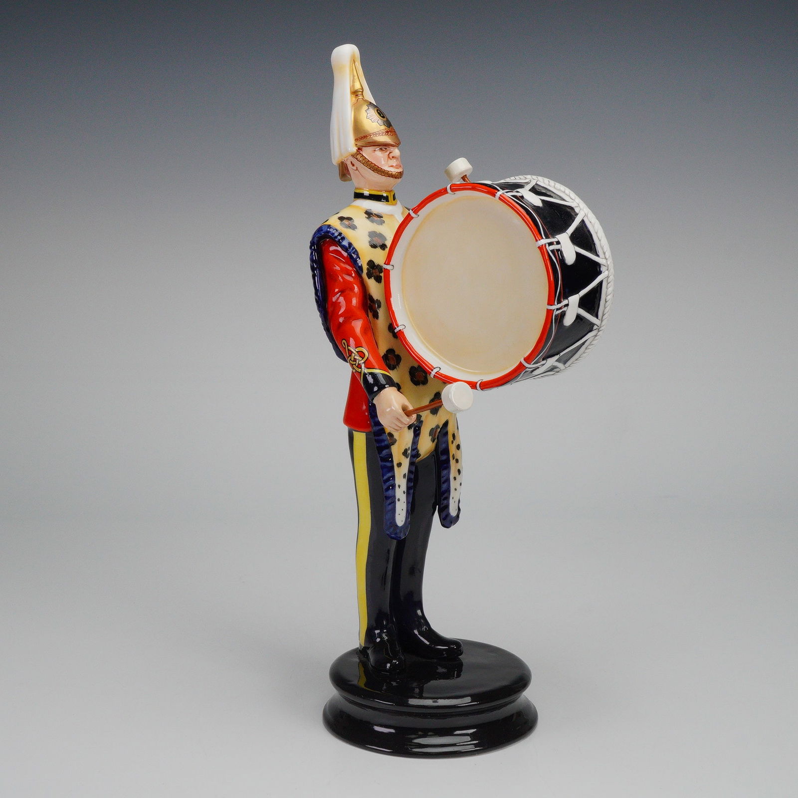 Michael Sutty Porcelain Sculpture, 1st Kings Dragoon Guards: Shown in full 1st Kings Dragoon Guards band uniform, the figure carries a large bass drum positioned across the front, with both drumsticks held in a playing stance. The uniform includes crested helme