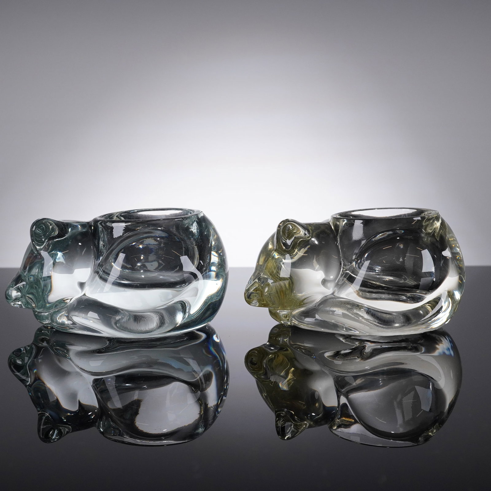 A Pair of Vintage Avon Glass Sleeping Cat Votive Holders: A pair of vintage molded glass candle holders produced by Avon, commonly known as the Sleeping Cat Votive Holders. Each holder is crafted from heavy, clear glass, stylized as a reclining cat figure wi