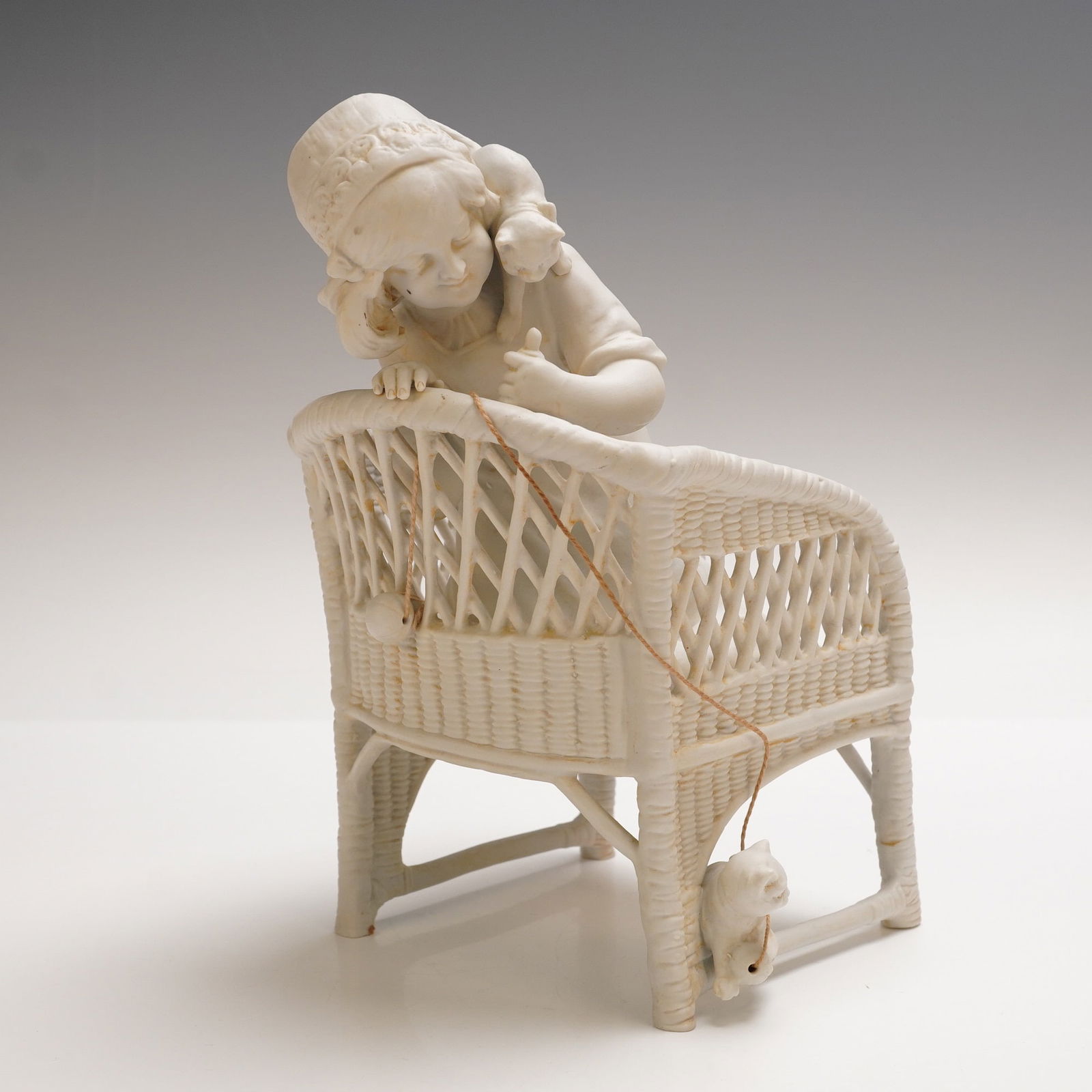 Bisque Porcelain Girl with Cats in Wicker Chair: Delightful 9-inch bisque porcelain figurine of young girl peering over the back of an intricately woven wicker chair. Cat perched on her shoulder with another grabbing at a ball of string. Piece is un