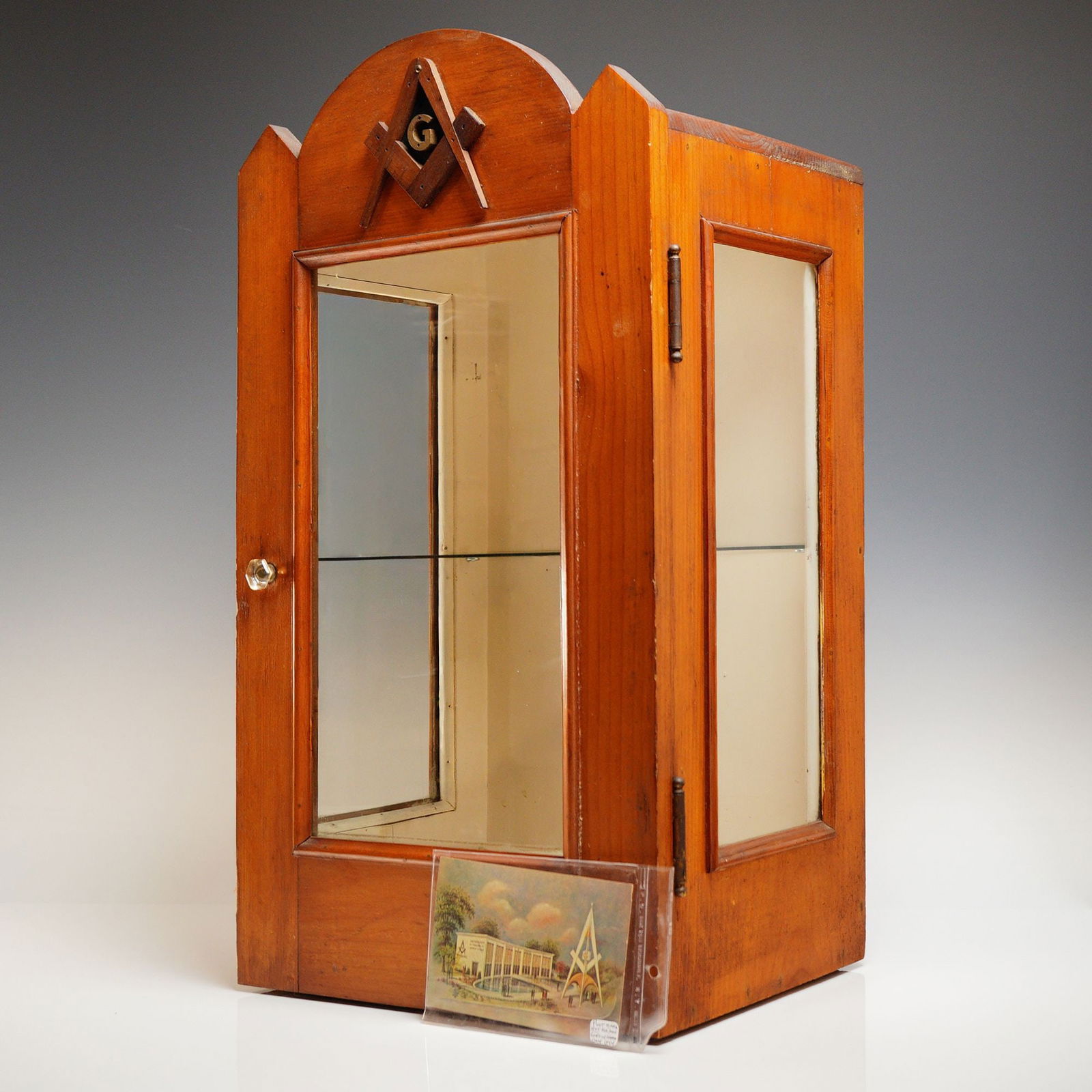 Vintage Masonic Display Case with Original Postcard: A vintage Masonic display case constructed of medium-stained pine or similar softwood, featuring an arched crest. The case is adorned with an applied dark-toned Masonic Square and Compasses emblem enc