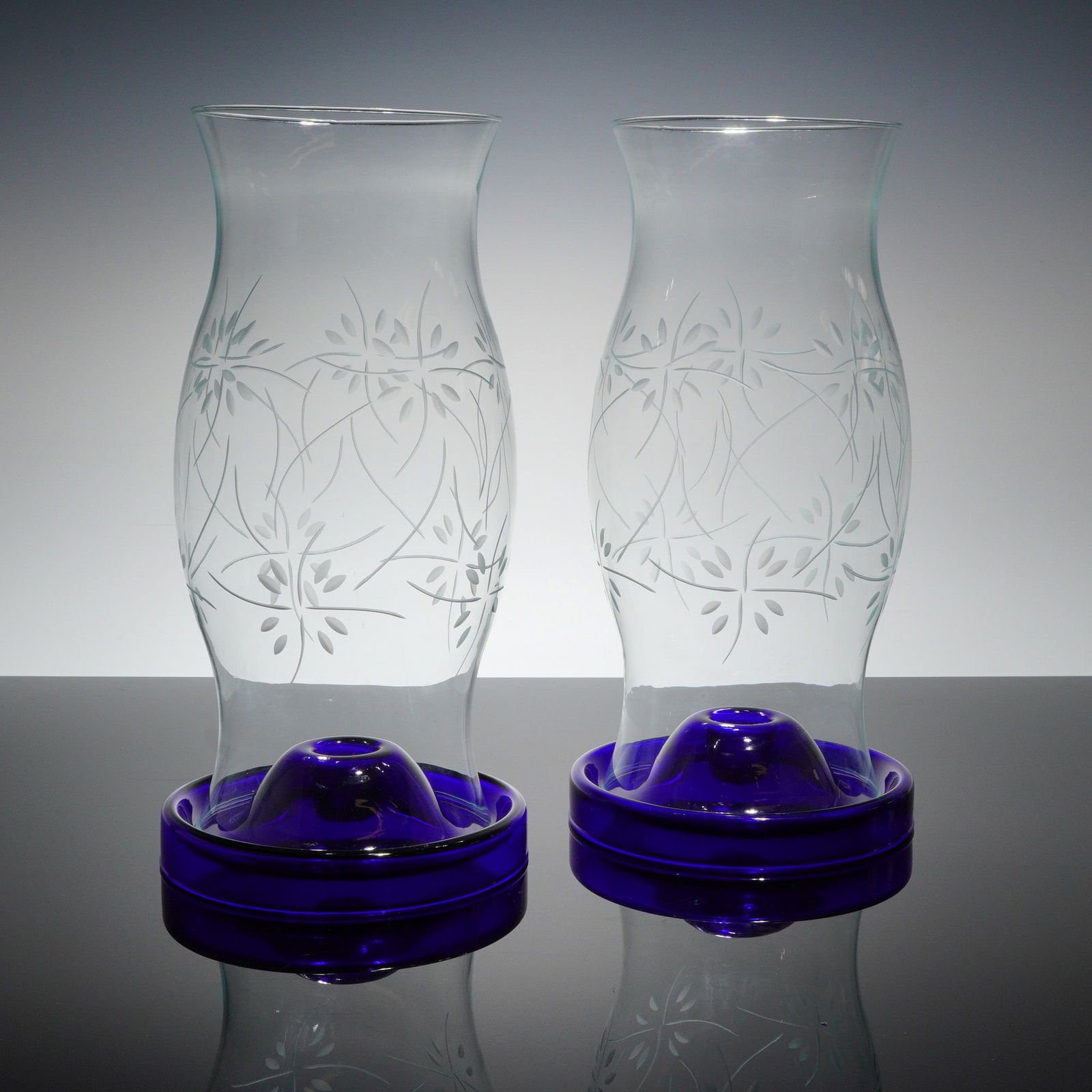 A Pair of Cobalt Blue and Clear Glass Hurricane Candle Lamps: This lot consists of a pair of contemporary hurricane candle lamps, presented as a 4-piece Glass candle holders set. Each lamp is constructed in two separable parts: a tall, slightly waisted clear gla