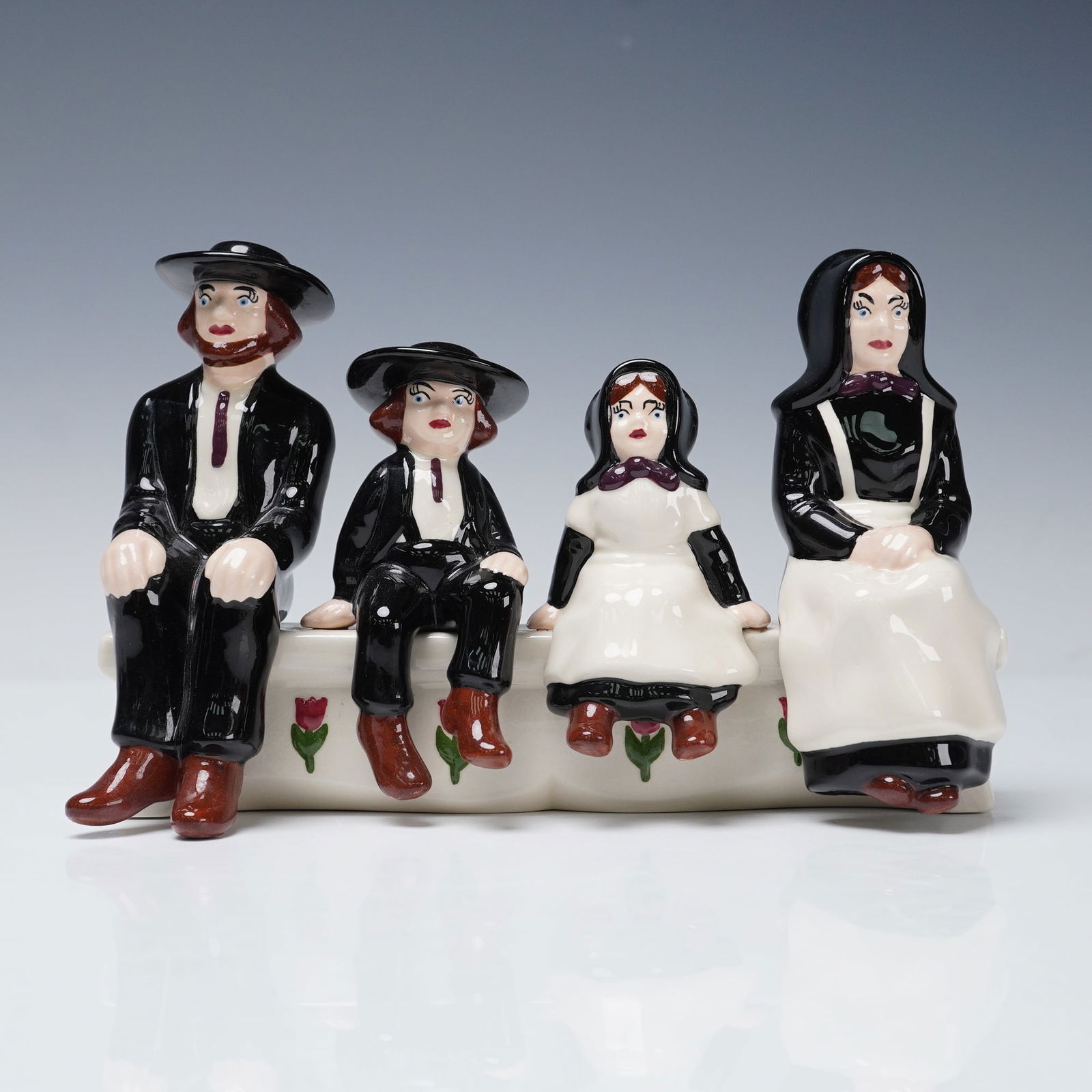Holland Mold Glazed Ceramic Amish Family Figurine: A mid 20th century glazed ceramic figurine by Holland Mold depicting an Amish family of four seated together on a tulip adorned bench. The grouping includes father, mother, son, and daughter, each dre