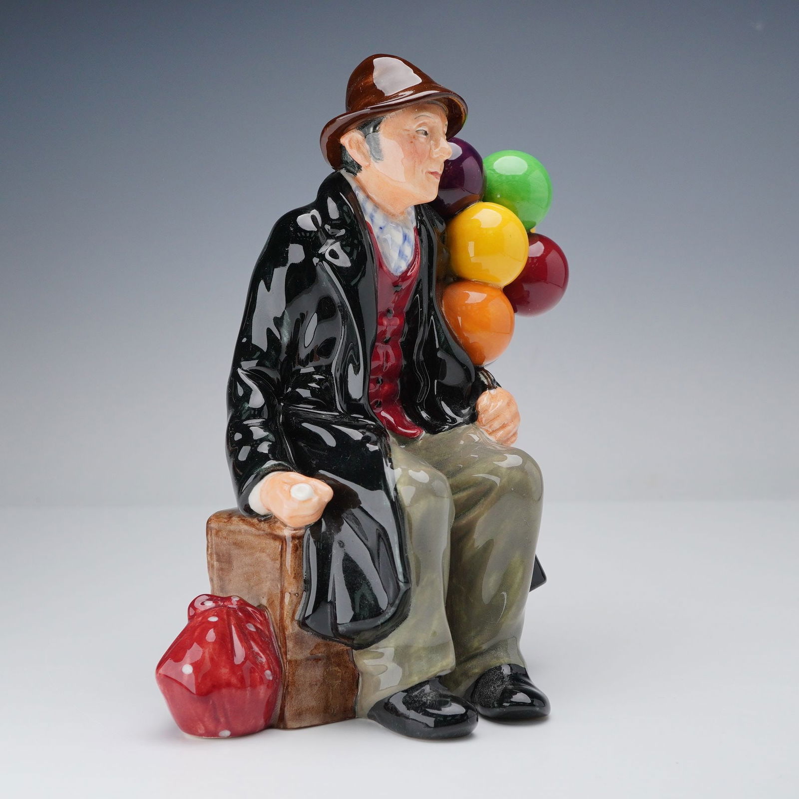 Royal Doulton Figurine, The Balloon Man HN1954: This Royal Doulton figurine titled The Balloon Man HN1954 features a charming scene of an elderly man sitting on a crate, holding a bundle of colorful balloons. The intricate detailing in his attire,