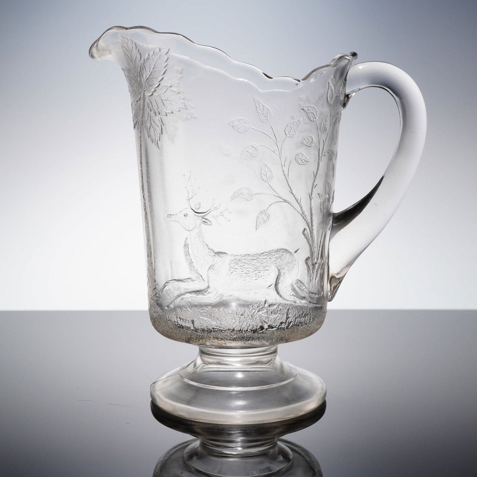 EAPG Deer and Oak Tree Water Pitcher: A colorless Early American Pressed Glass (EAPG) water pitcher. The vessel features a tall, slightly tapered body mounted upon a pronounced, tiered pedestal base. The decoration is extensive and geomet