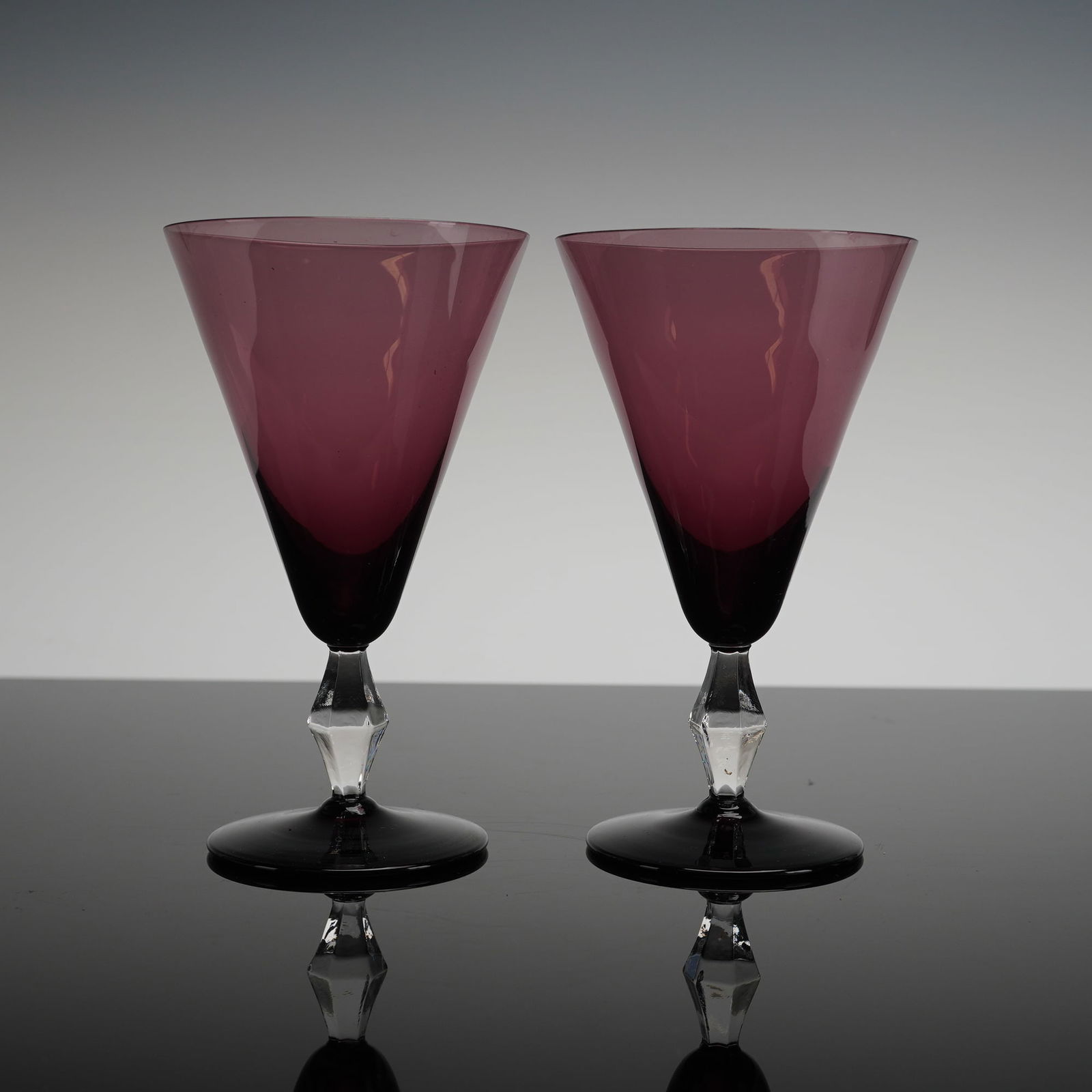 A Pair of Purple Amethyst Glass Martini Glasses: This 2pc set features a modern aesthetics. Each glass has a wide, inverted conical bowl constructed from translucent purple glass. The color blends into a darker, near-opaque black towards the base of