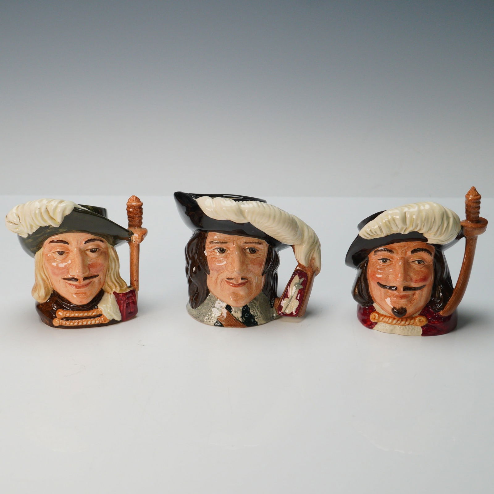3pc Royal Doulton Small Character Jugs, Musketeers: Trio of glazed ceramic character jugs depicting the musketeers Athos, Porthos, and D'Artagnan, each wearing a plumed hat with the handle modeled as a sword hilt. All are finished in vibrant coloration