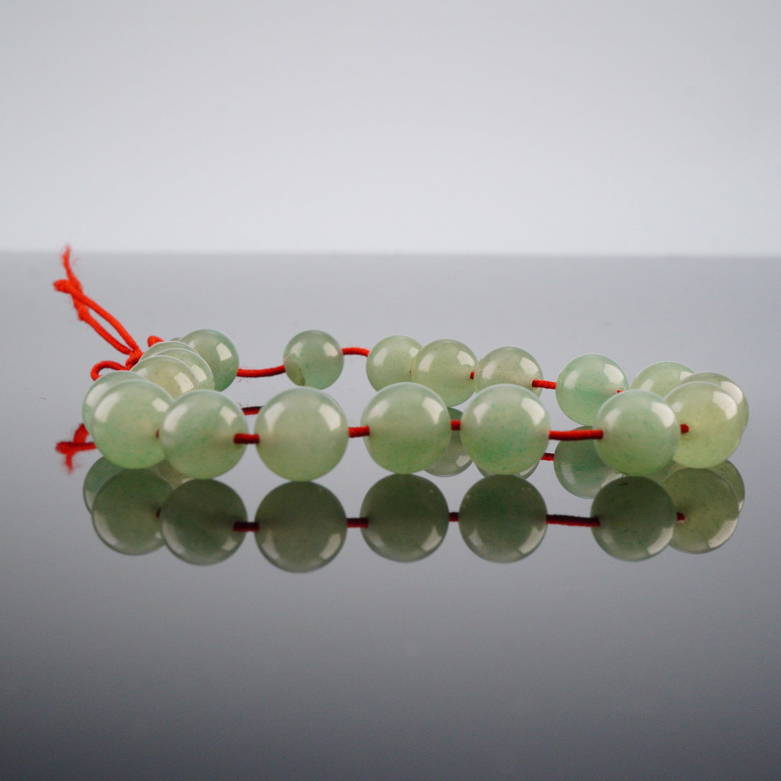 Natural Green Jade Beaded Bracelet: Natural green jade beaded bracelet, comprising polished, spherical beads, strung on a red textile cord. The beads exhibit a translucent to semi-translucent light green hue, with a smooth, lustrous sur
