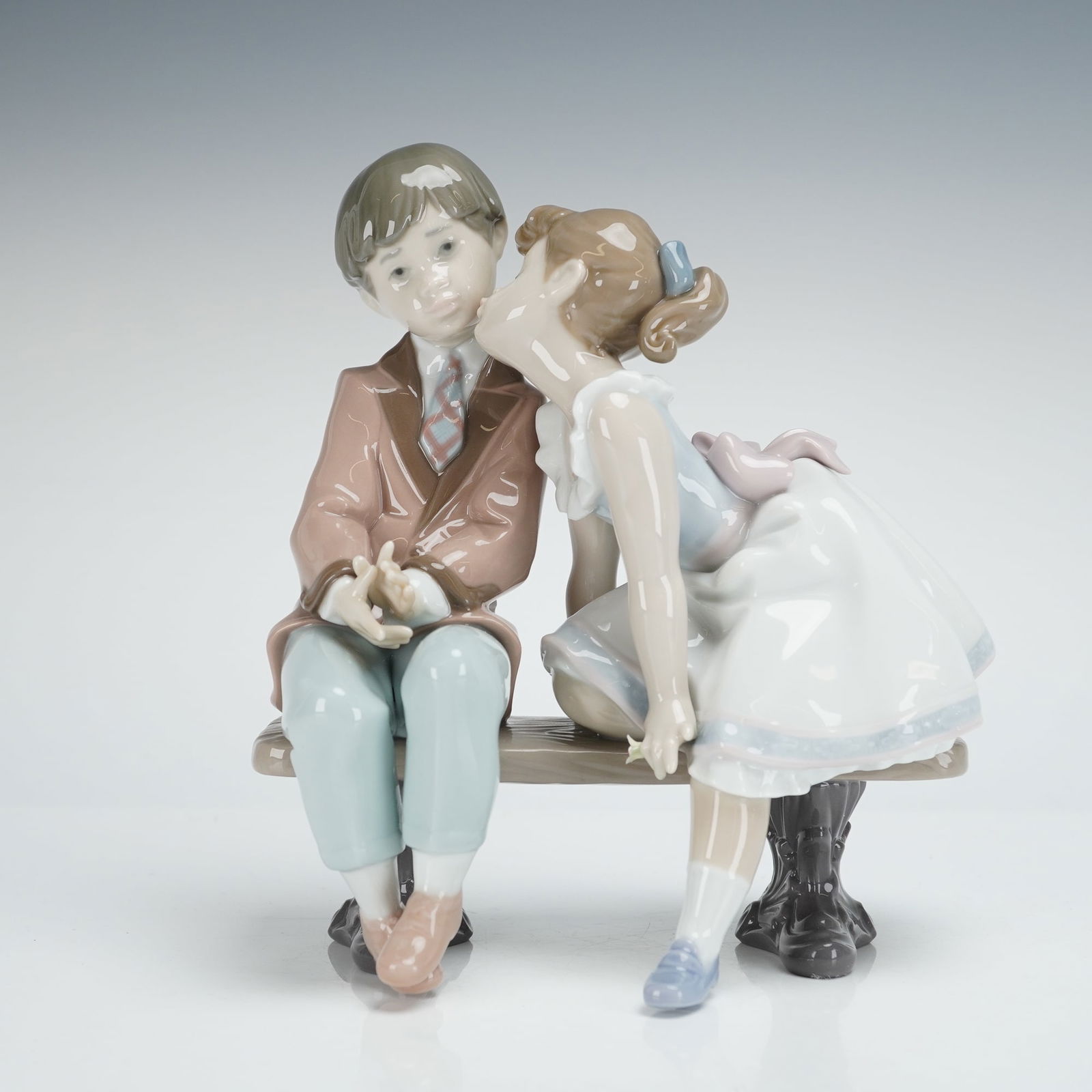 Lladro Porcelain Figurine Ten and Growing 01007635: The figurine depicts a sentimental scene of a young boy and girl seated on a park bench, with the girl leaning in to kiss the boy's cheek. The figures are rendered in subtle pastel coloring, including