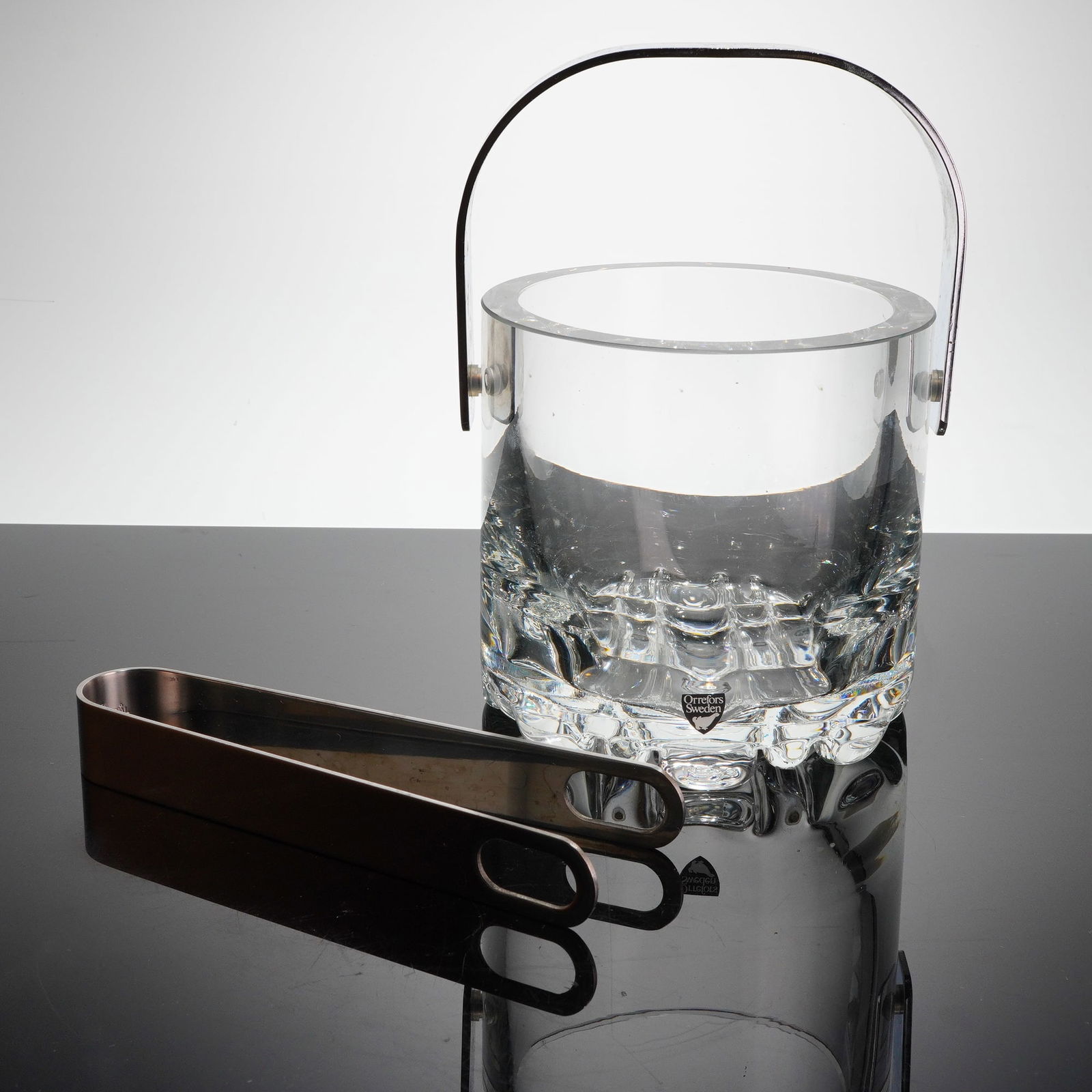 Orrefors Erik Patern Crystal Ice Bucket and Tongs: A two-piece set featuring the Orrefors Erik patern ice bucket, often atributed to designer Olle Alberius. The heavy, cylindrical vessel is constructed of clear lead crystal or thick glass, consistent