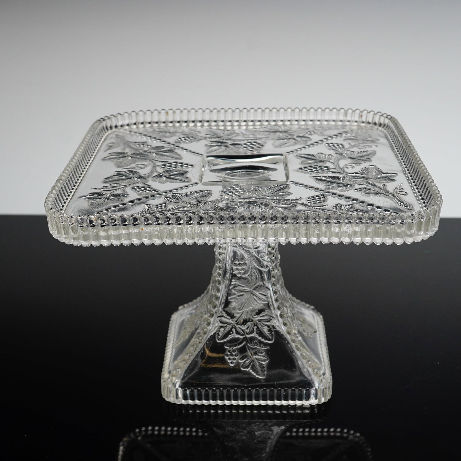 U.S. Glass EAPG Square Cake Stand, Beaded Grape: The item features a square plate resting on a tapering pedestal base. Both the plate surface and the base are molded with a design of grapevines. These motifs are separated by diagonal beaded borders